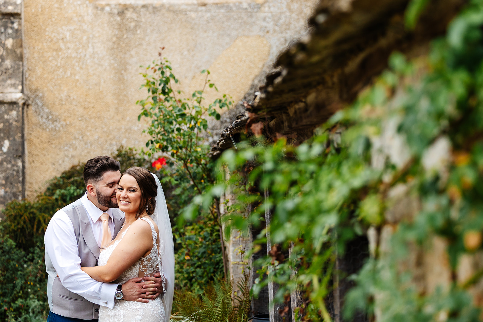 Couple in grounds of Caswell House with flowers and greenery in the background, Groom kisses Bride on the cheek