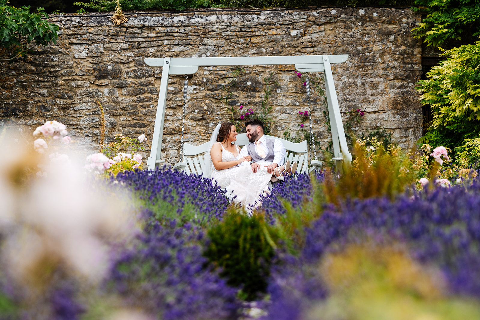 Couple sit on white wooden swing in the grounds of Caswell House with an exposed brick wall in the background, Lavender and flowers surround them 