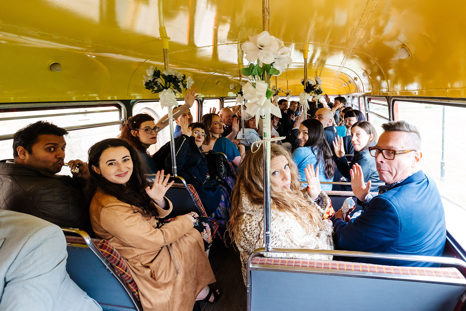 A group of wedding guests smiling at the camera and waving on the way to a wedding in a double decker bus.