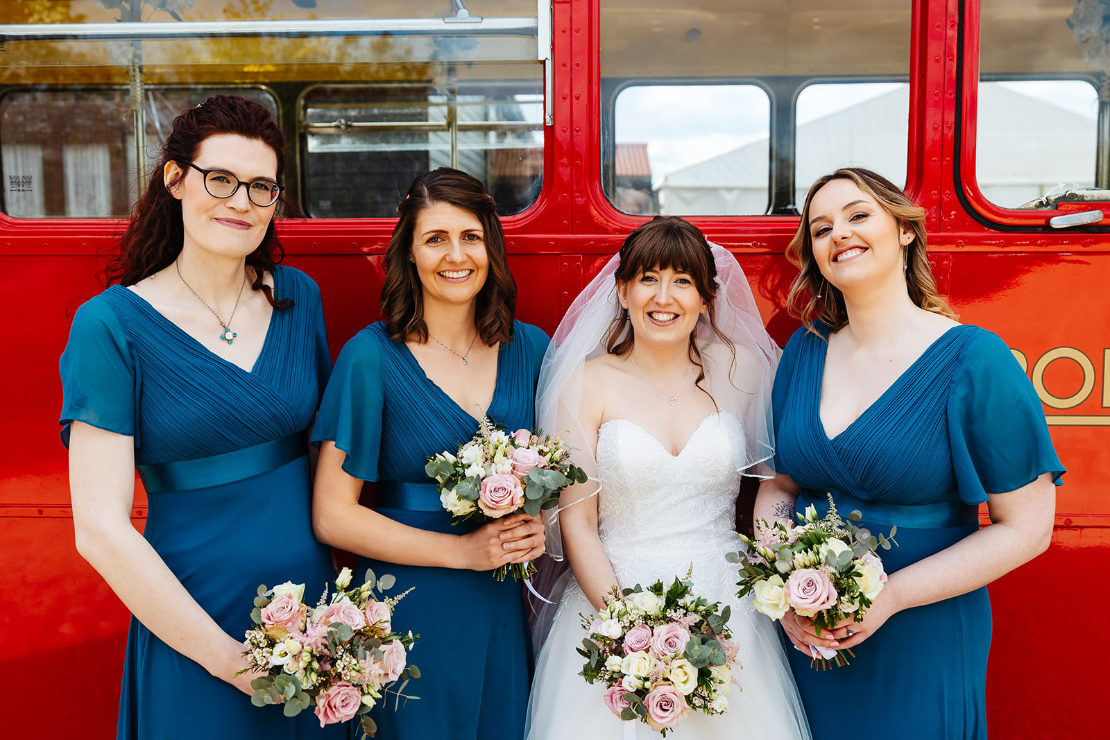 A bride and three bridesmaids standing outside a red double decker bus smiling and holding flowers