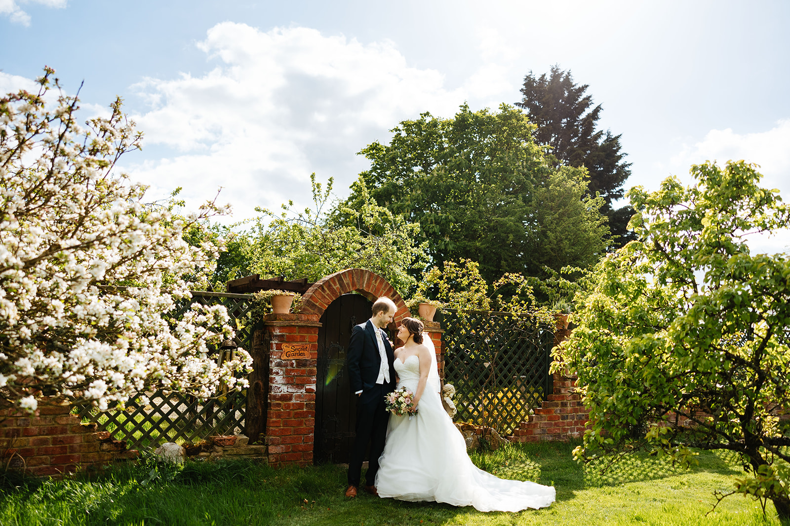 A couple outside enjoying their wedding day. The groom is in a blue suit with brown shoes on and the bride is in a long, flowing off the shoulder dress with a veil
