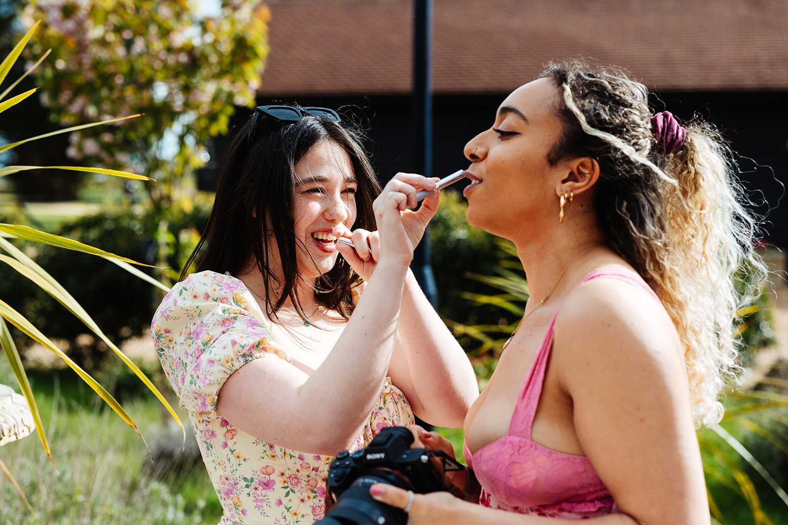 Two wedding guests outside putting on lipstick and smiling in the sunshine