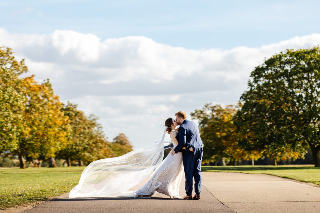 Bride and groom standing in the middle of the driveway at bassmead manor barns. They're surrounded by green trees, and the brides veil floats in the breeze.