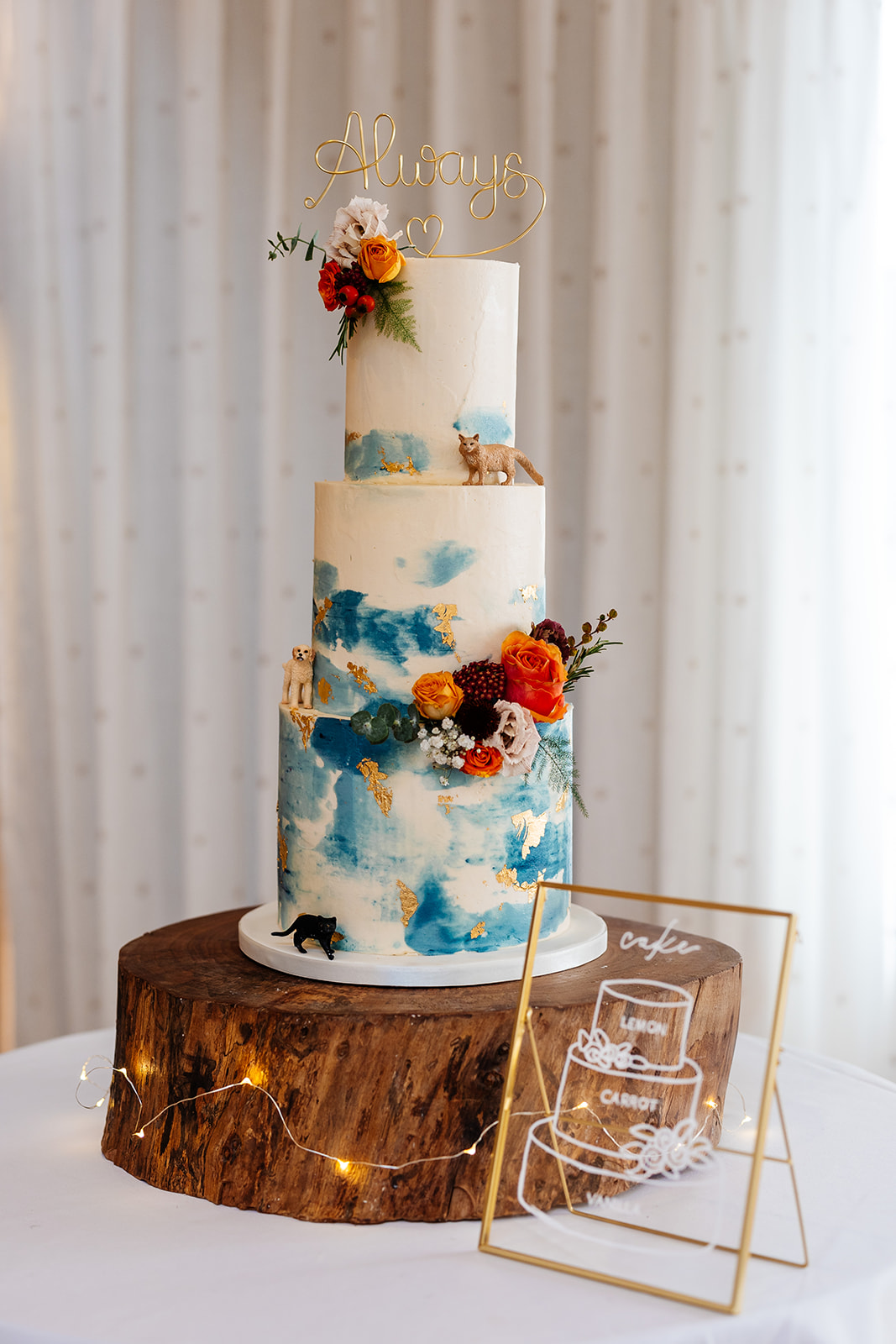 A three tiered white wedding cake with blue decorations and orange and yellow flowers on the cake. The cake is standing on a wooden log piece.