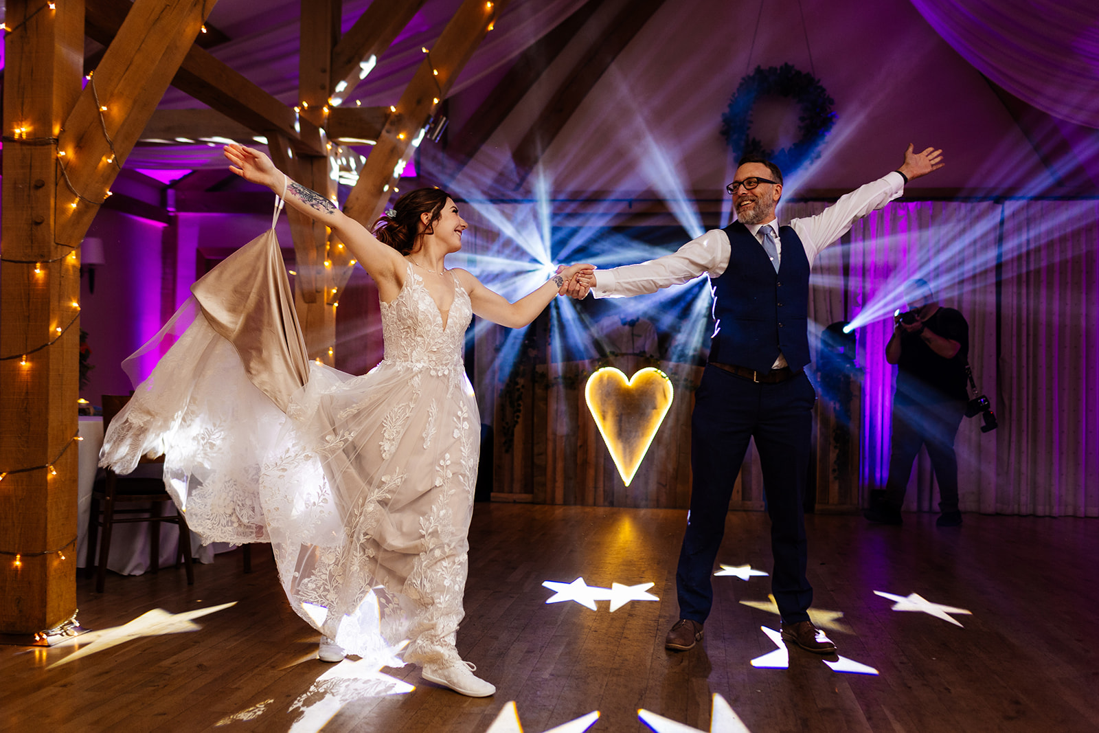 The Father of the bride and the bride are in an open arm position dancing on the dance floor with lights all around them