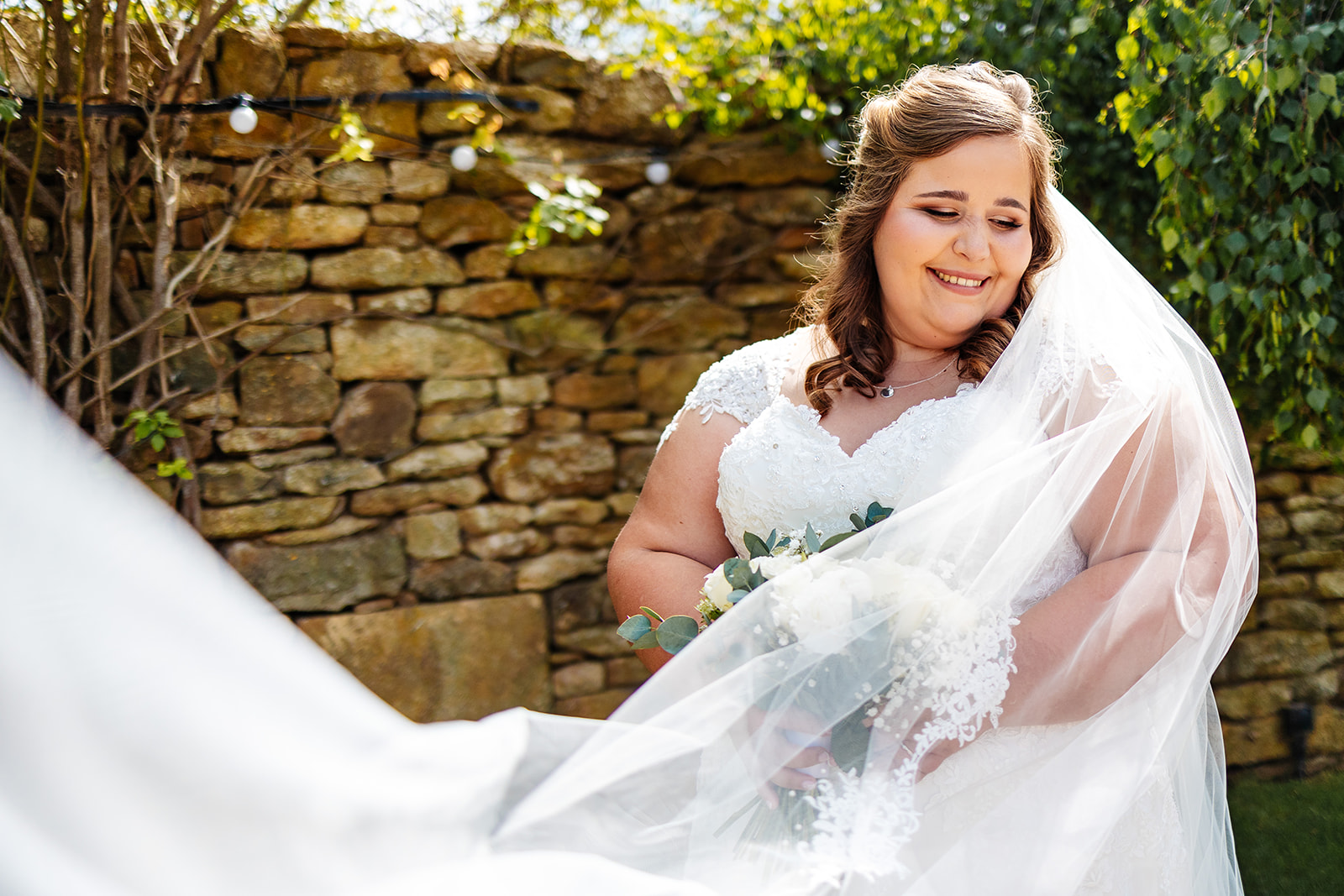 A bride outside with her veil floating in the air smiling holding a bunch of flowers