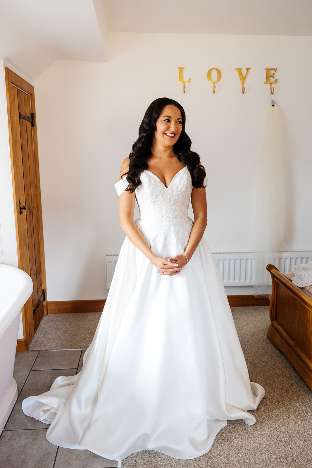 A bride in her wedding dress. The dress is off the shoulder and her brunette hair is wavy hanging over her shoulders