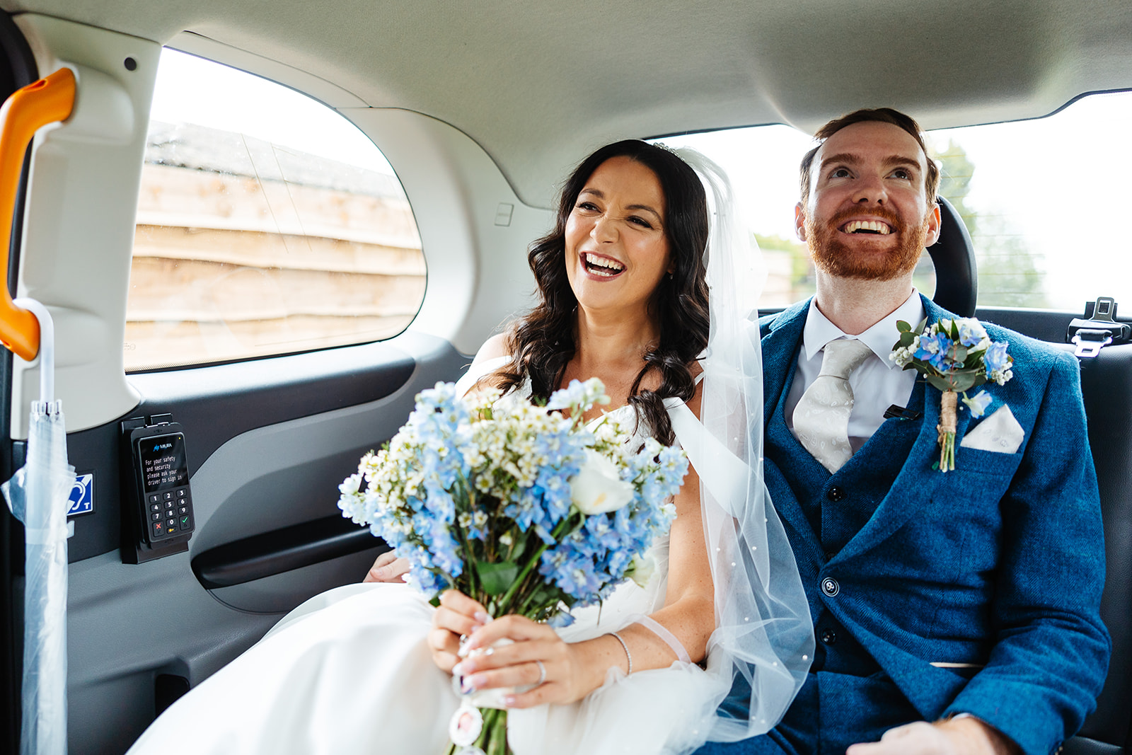 A bride and groom in a taxi after their wedding. The groom is dressed in a navy suit and the bride is holding a bouquet of pale blue and white flowers