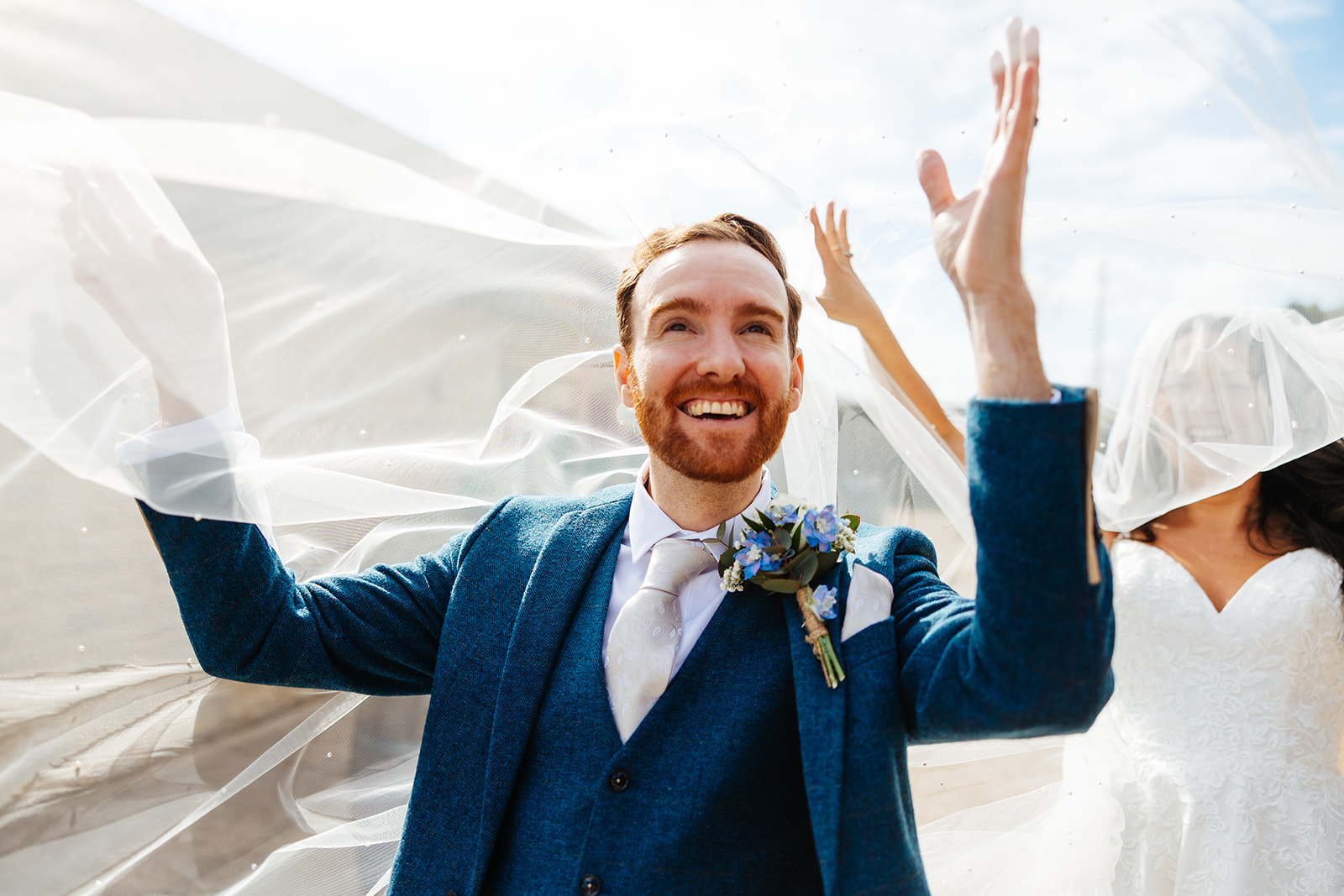 A groom in a navy suit underneath his wife's veil as it floats around in the wind