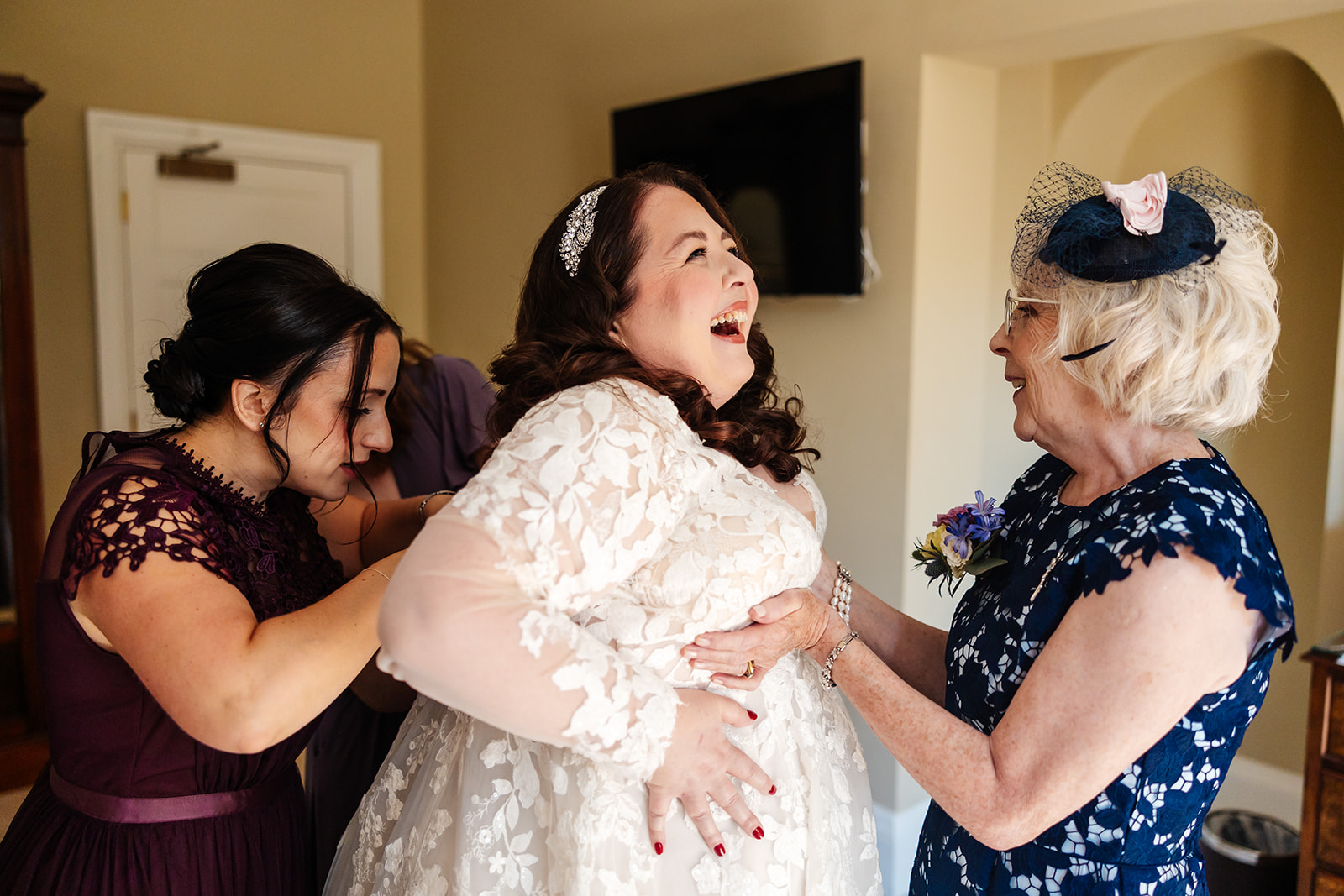 A Bride in her wedding dress being helped by her Mother and bridesmaid and they are all smiling