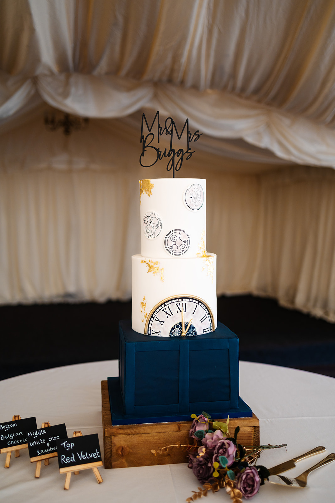 A three tier wedding cake with the top two tiers in white, decorated with a number of clocks on them and gold leaf and a blue bottom tier.