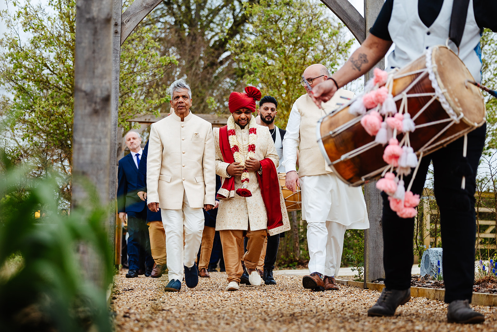 A groom, his father and his father in law walking on gravel towards their ceremony. There is a drummer in front who is playing the drum.