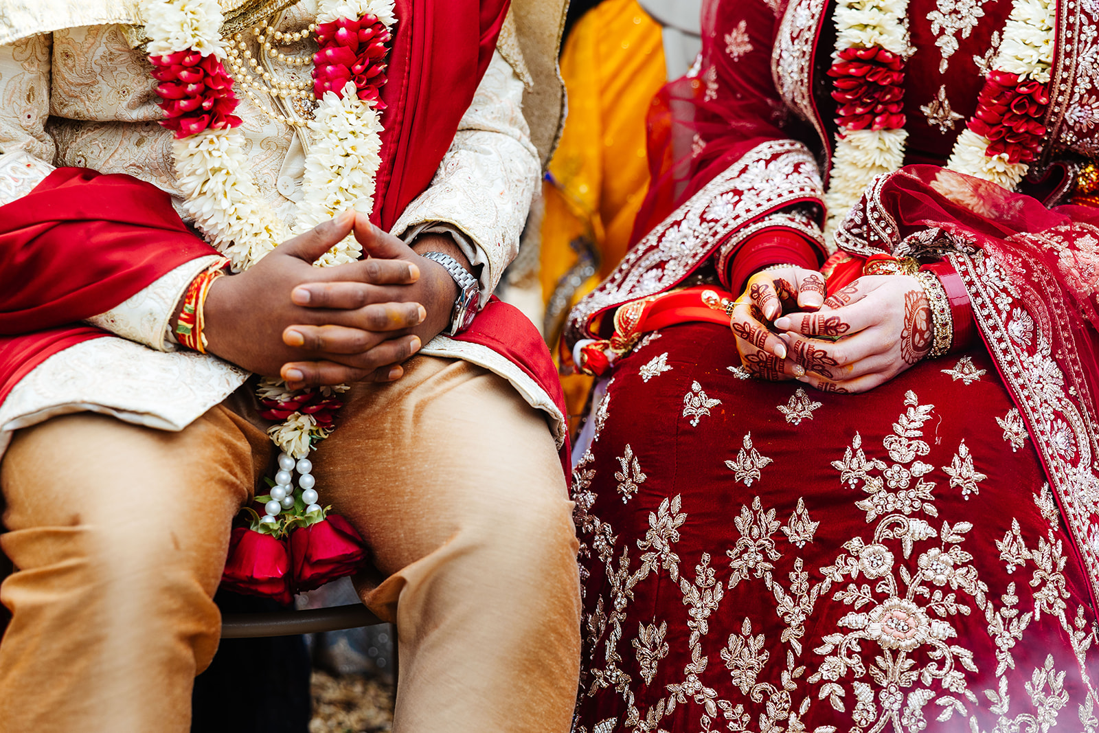 A bride and grooms hands in their lap. They are wearing golden and maroon outfits and the brides hand has henna on.