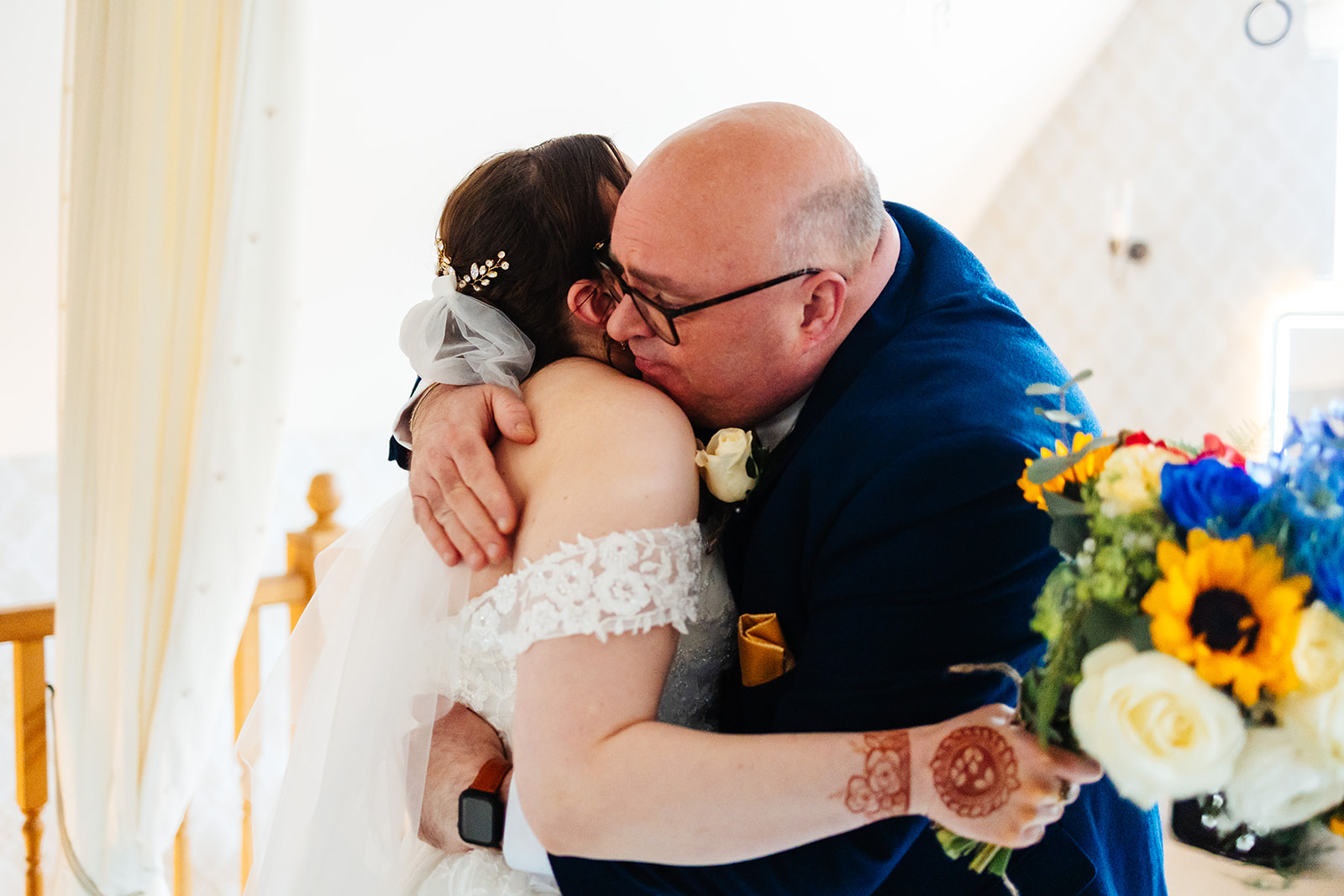 A father in a navy suit is hugging his daughter in a bridal dress as he sees her in her dress for the first time