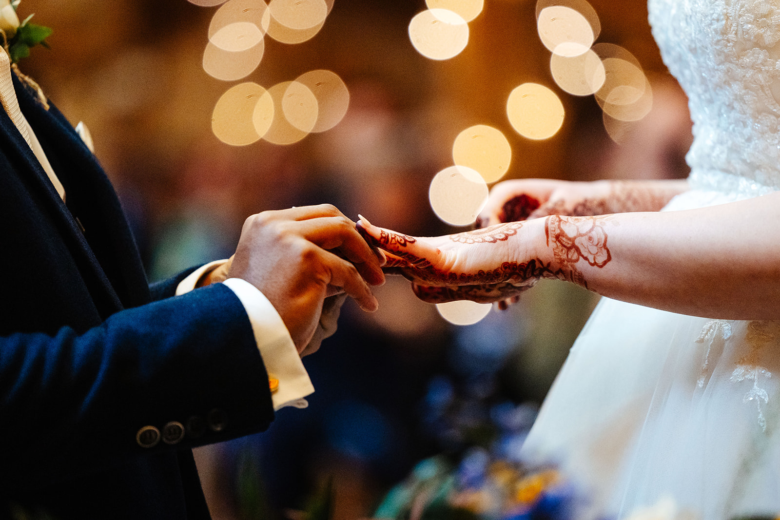 A bride and groom's hands, the bride is covered in beautiful henna tattoos on her hands as they exchange rings