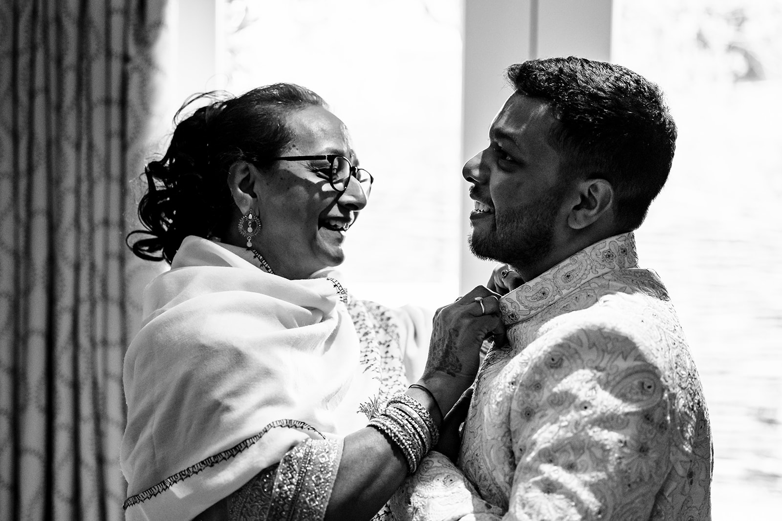 A groom and his mother. His mother is doing the top button of his shirt up and they are both smiling.

This photo is black and white