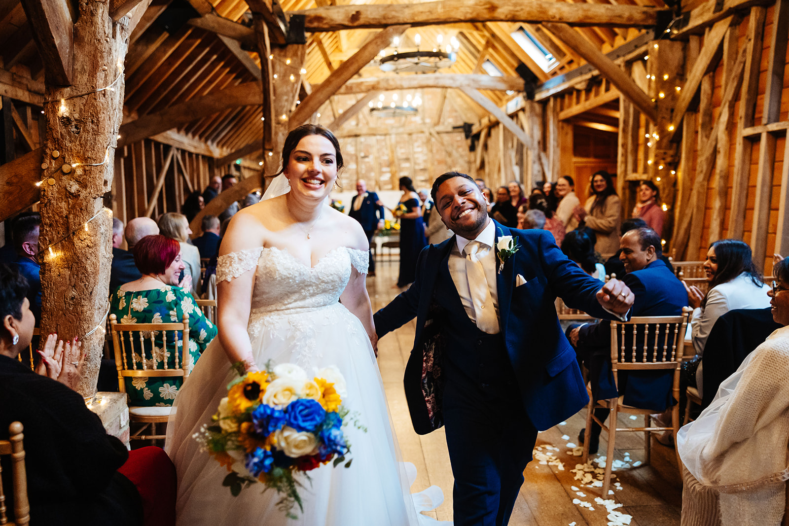 A newly married couple smiling walking down the aisle of a wooden barn and are gleefully walking down the aisle surrounded by loved ones.