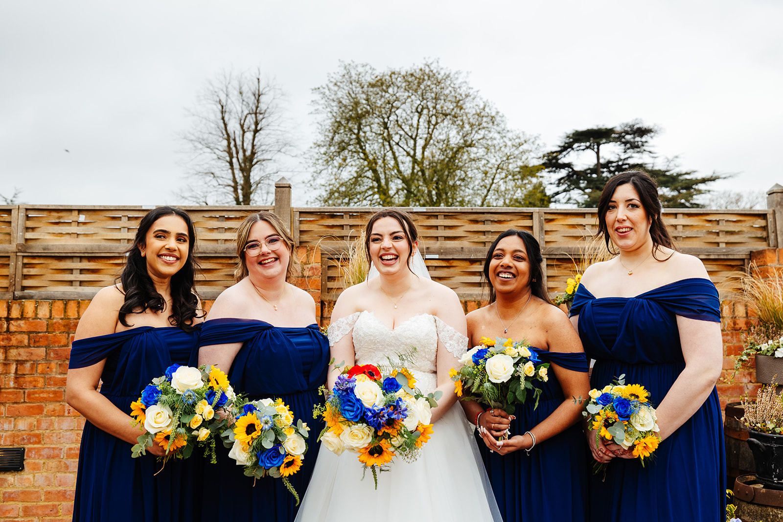 A bride in a white dress and her four bridesmaids in navy blue dresses holding a bouquet of blue flowers and sunflowers and are smiling. They are outside and behind a wooden fence
