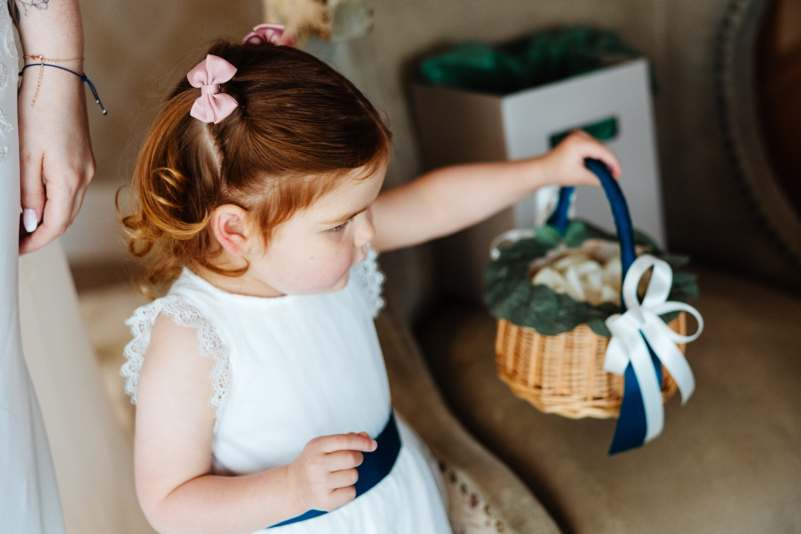 A little girl with ginger hair and half of it tied up with a little pink bow, wearing a white dress and holding a basket of petals.