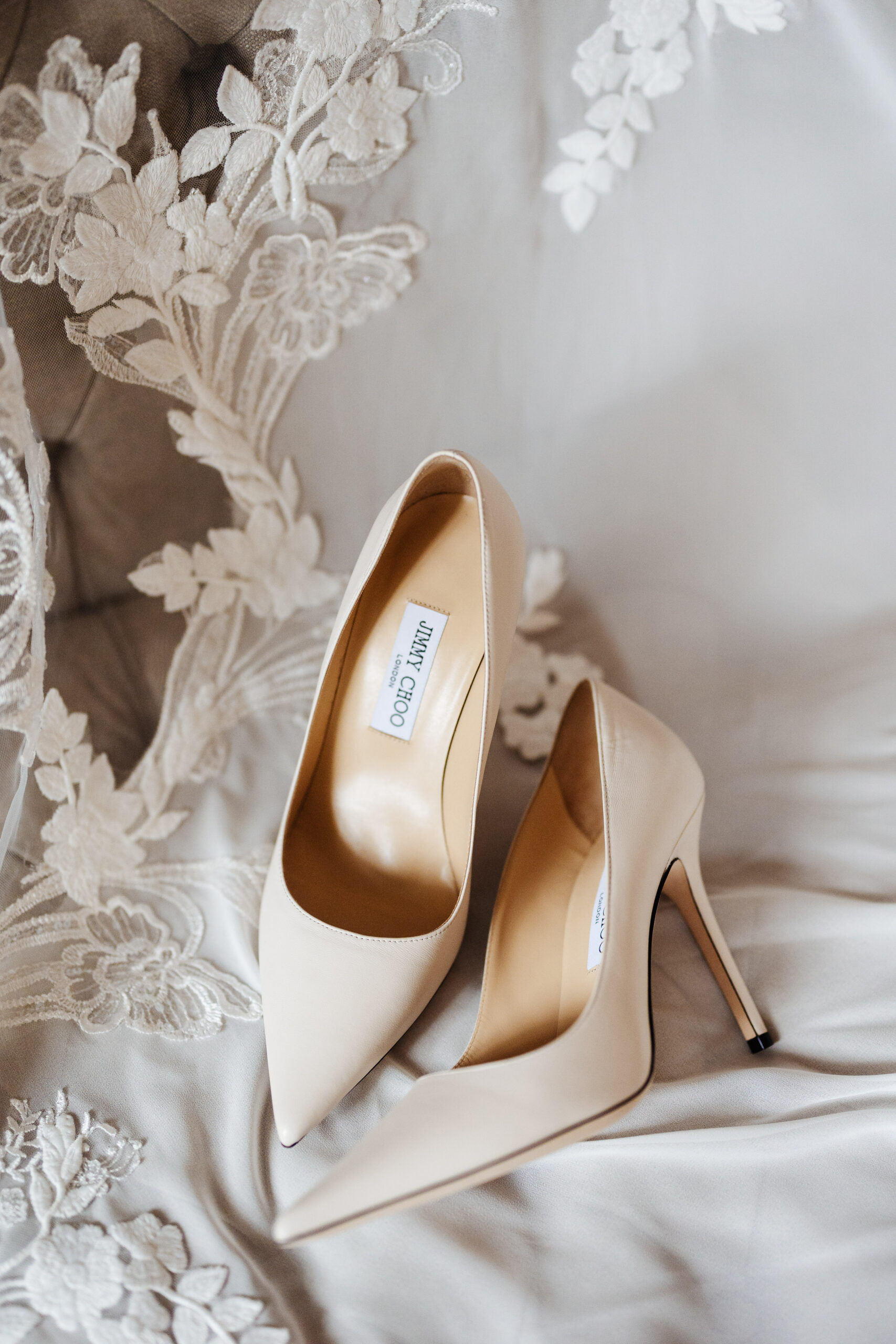 A pair of cream Jimmy Choo shoes on part of a white laced wedding dress.