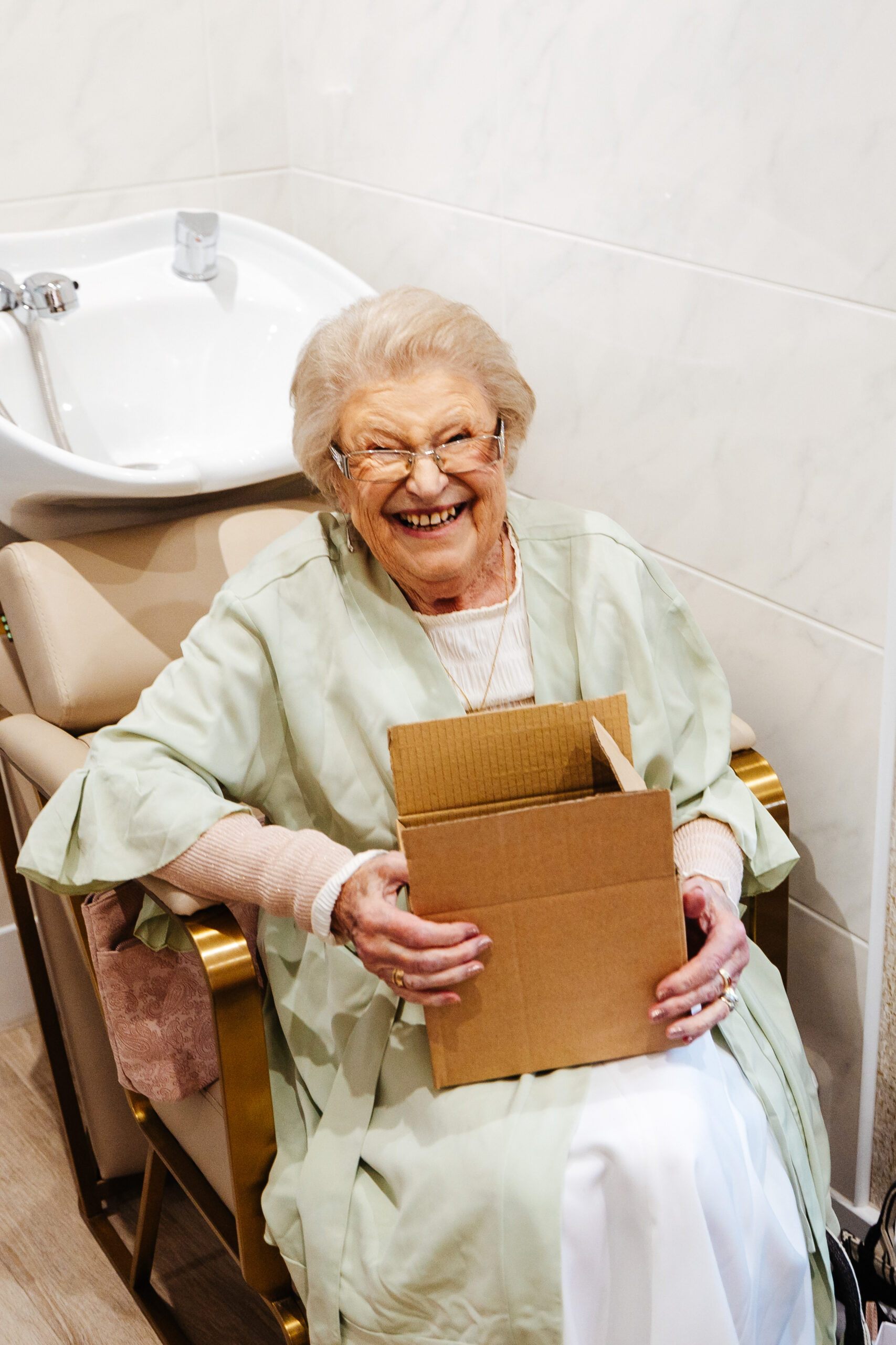 An older lady with a huge smile on her face sitting in a chair holding a wooden box.