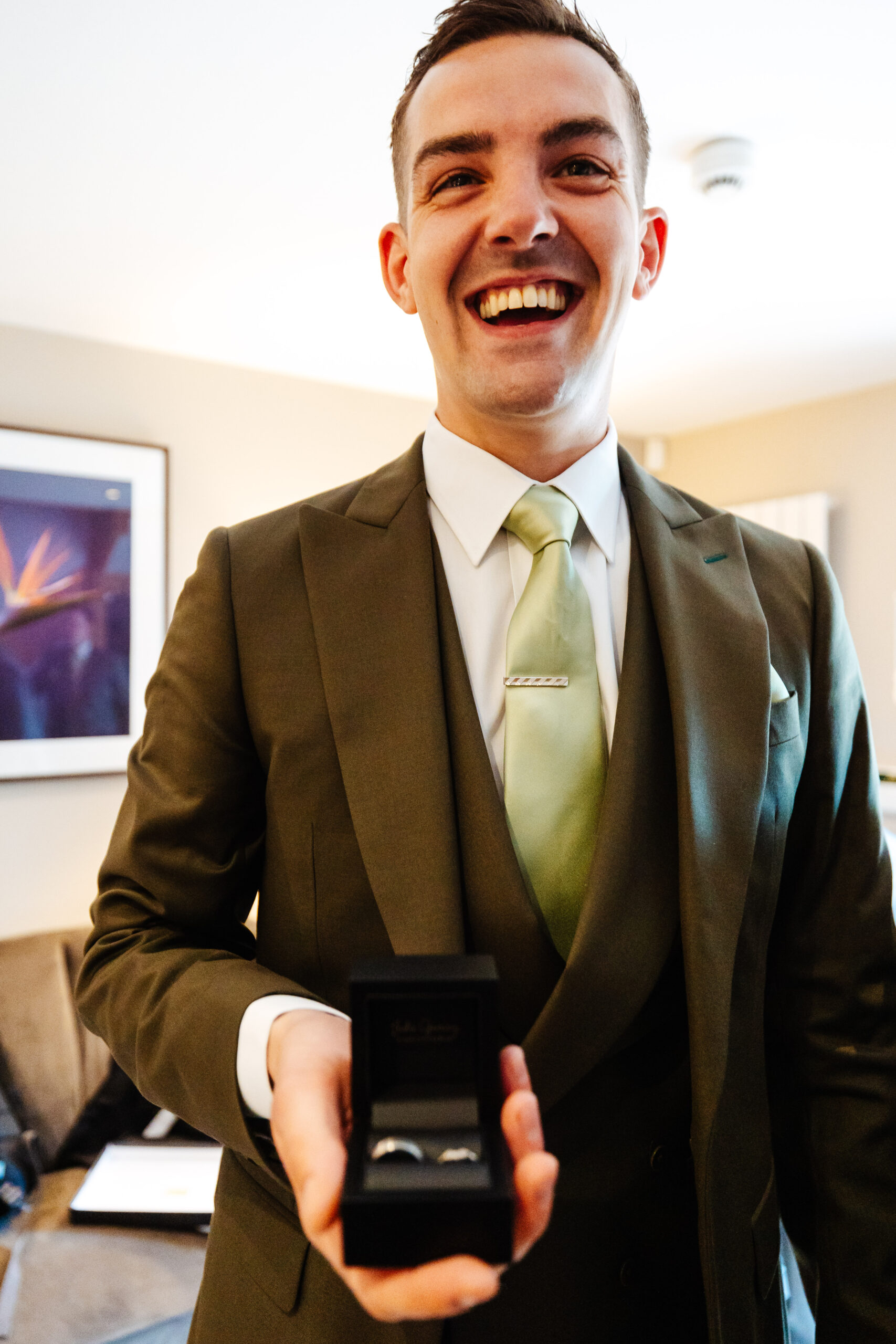 A man in a green suit with a huge smile on his face holding a ring box with a ring inside it.