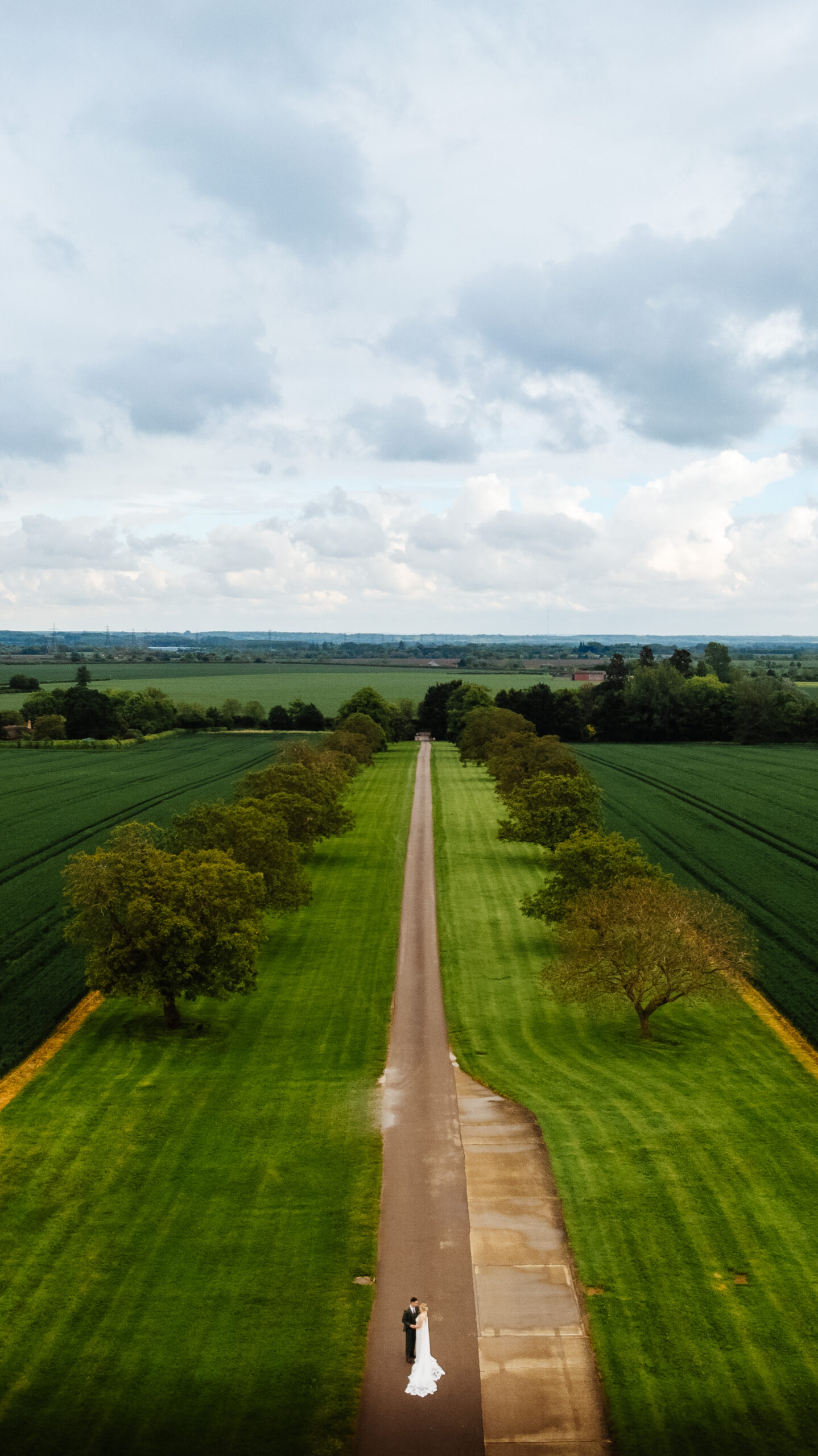 A large green field with a track running down the middle and a bride and groom in the middle of it all. The image is taken from high above.