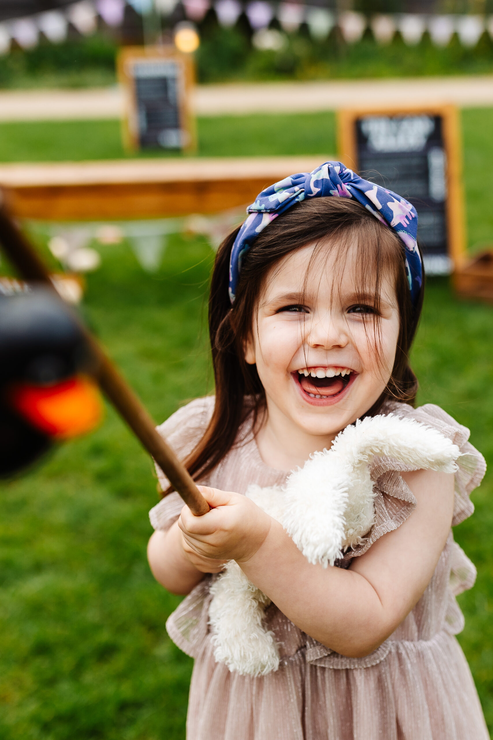 A little girl with brown hair a pink dress smiling at the camera. She is holding a stick as part of a garden game.