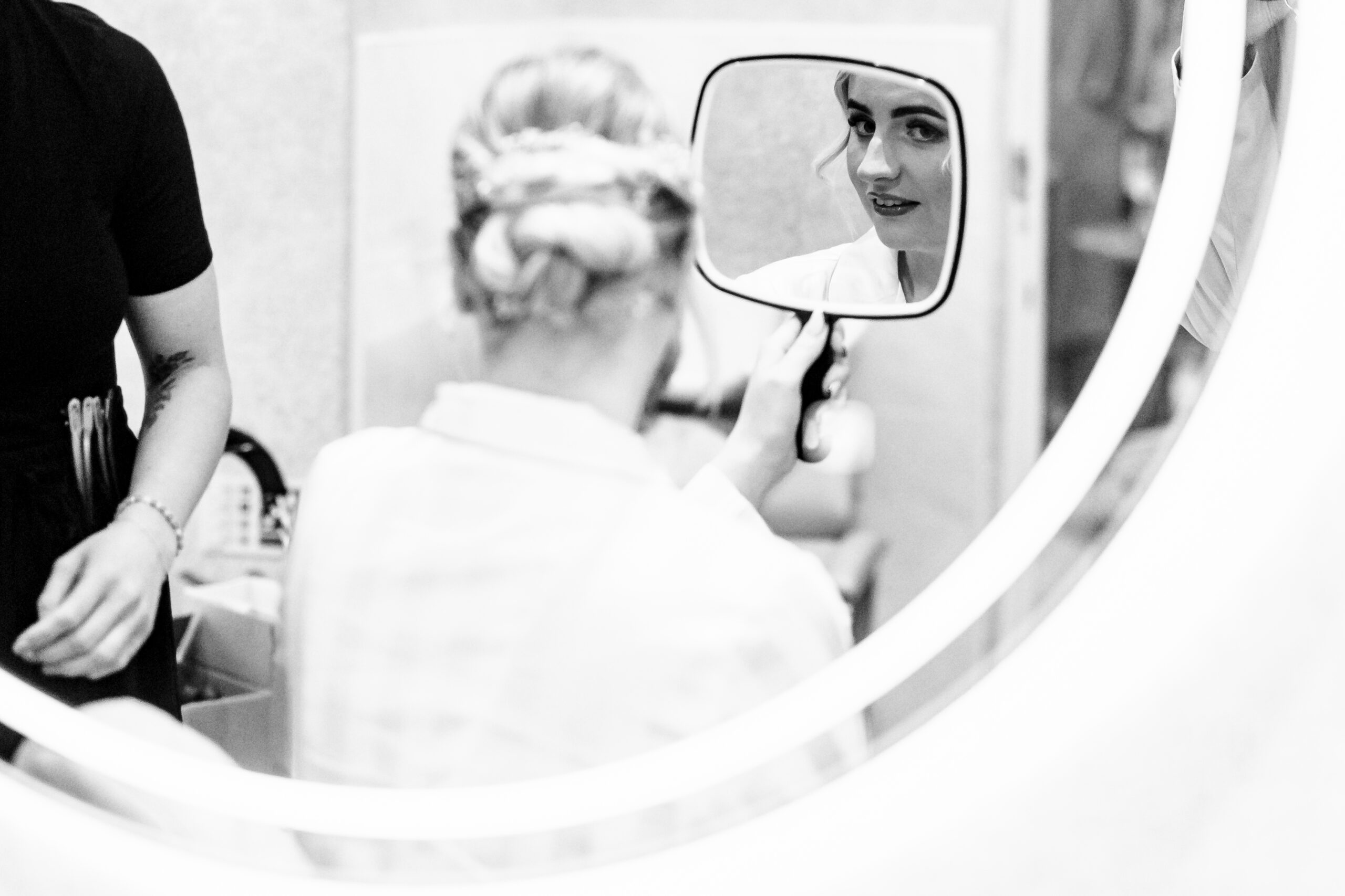 A black and white image of a bride in white silk pajamas looking at herself in a hand held mirror and smiling.