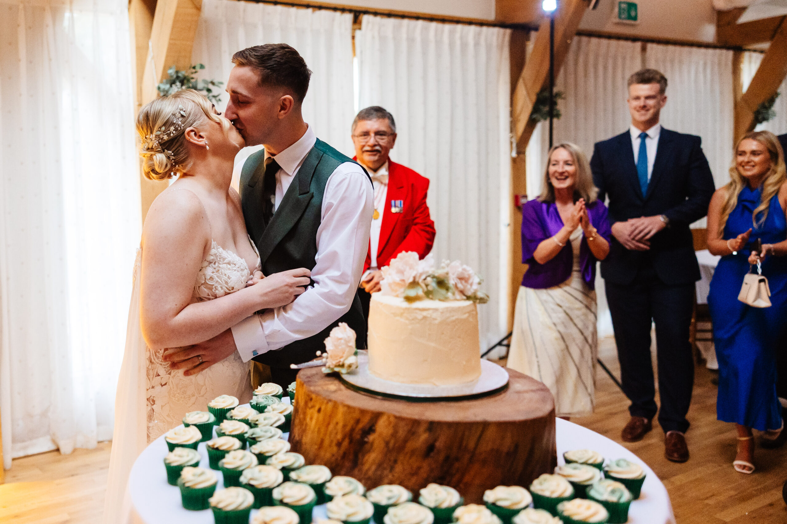 A bride and groom kissing in front of their wedding cake. The cake is one layered with cream frosting and beige flowers on top.