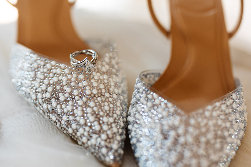 A pair of white, jewell encrusted high heels. A wedding ring is balanced on the shoe.