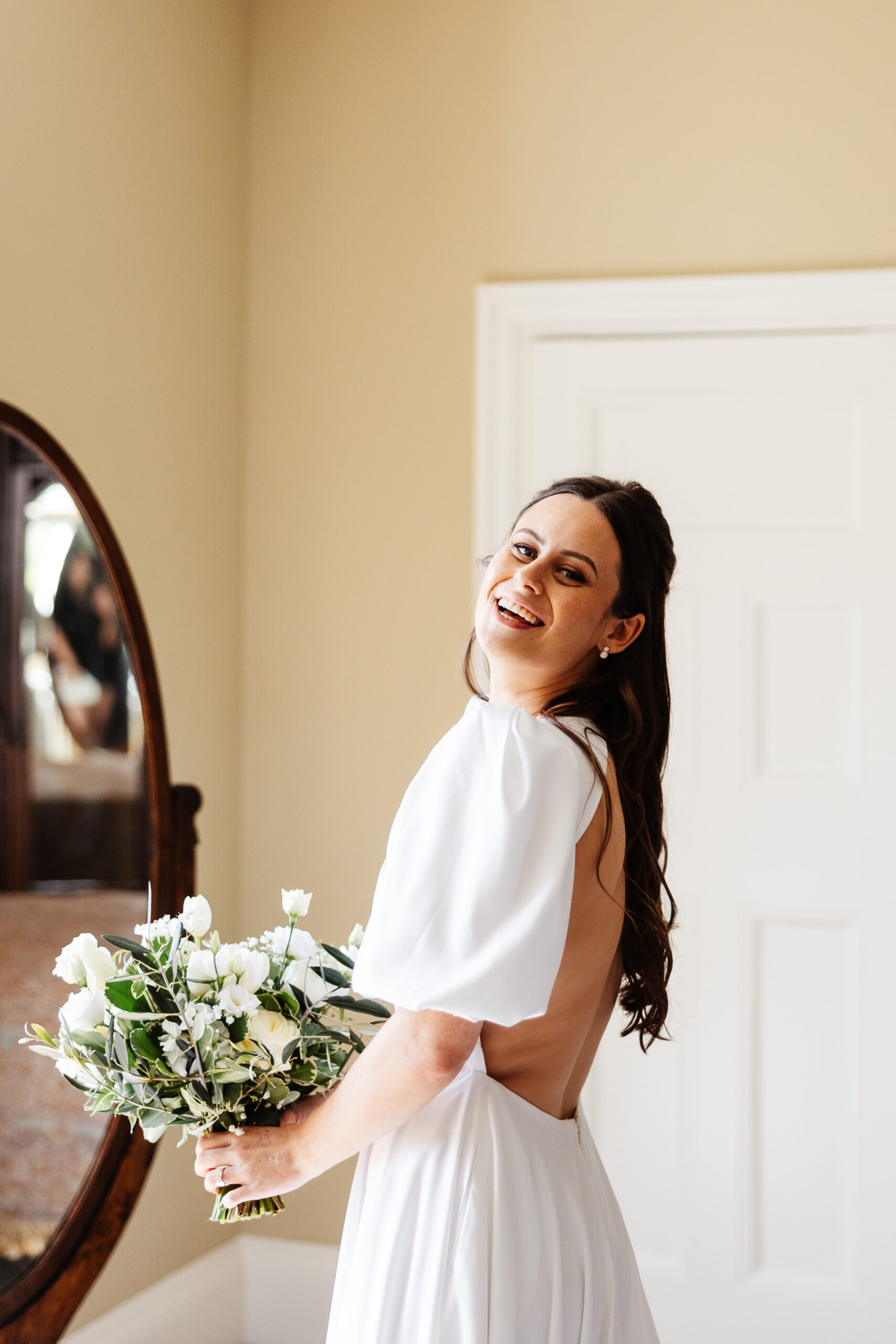 A bride in her white backless wedding dress, smiling at the camera holding a bunch of white flowers.