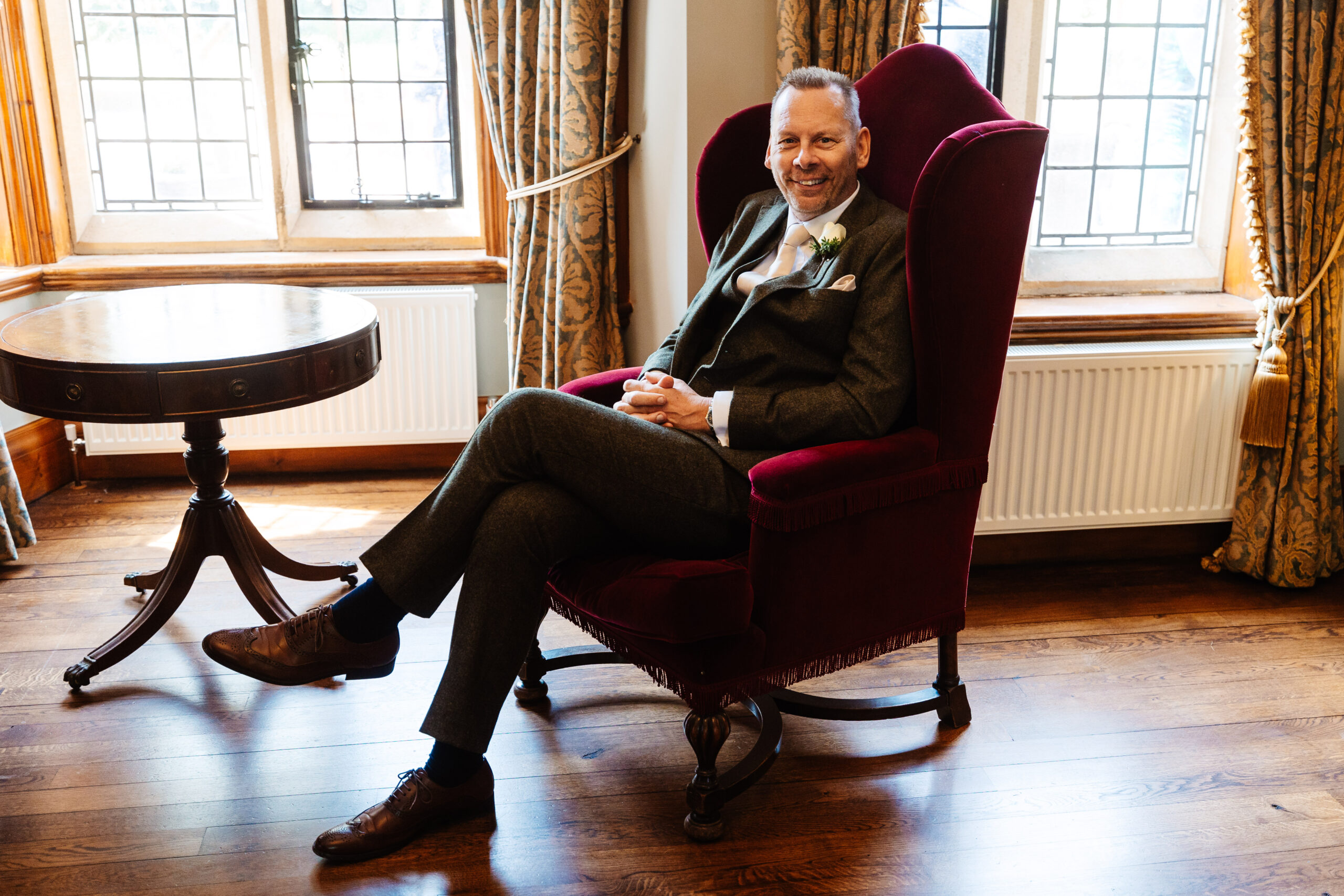 A man in a dark green tweed suit with his legs crossed sitting in a formal chair.