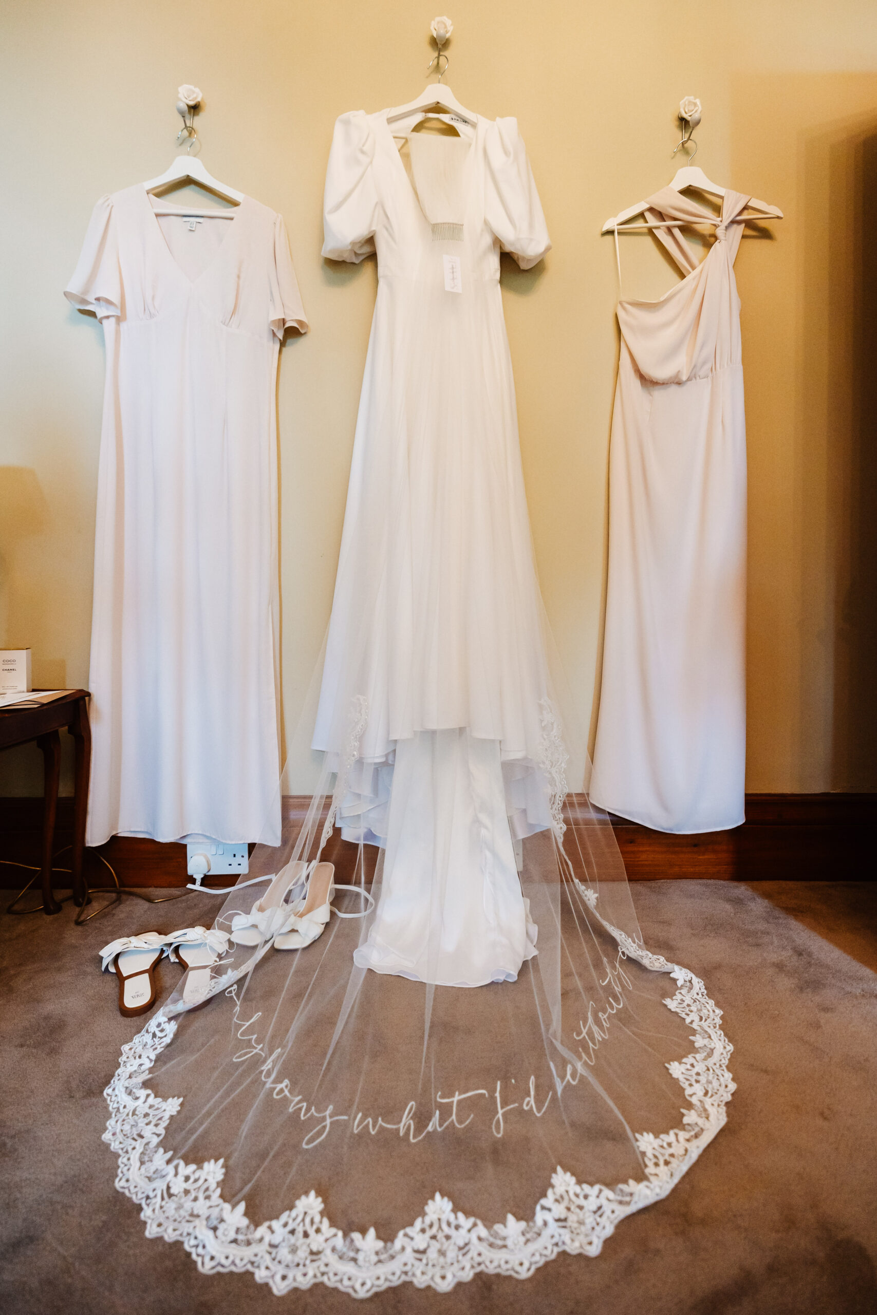 Three white dresses hanging up on a wall. One is a wedding dress with puffed sleeves and the other two are long, flowing dresses, one of them with an off the shoulder tie.