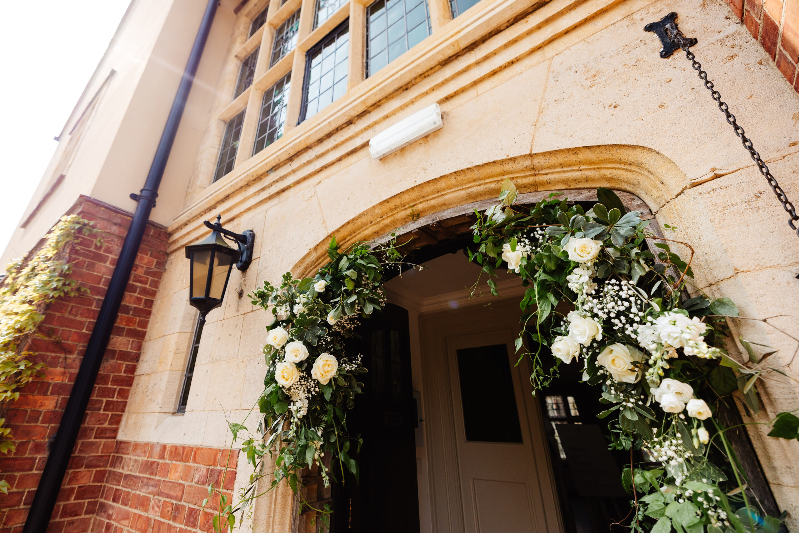 An archway of an old stone building and hanging from the arch is white roses, greenery and little white buds.