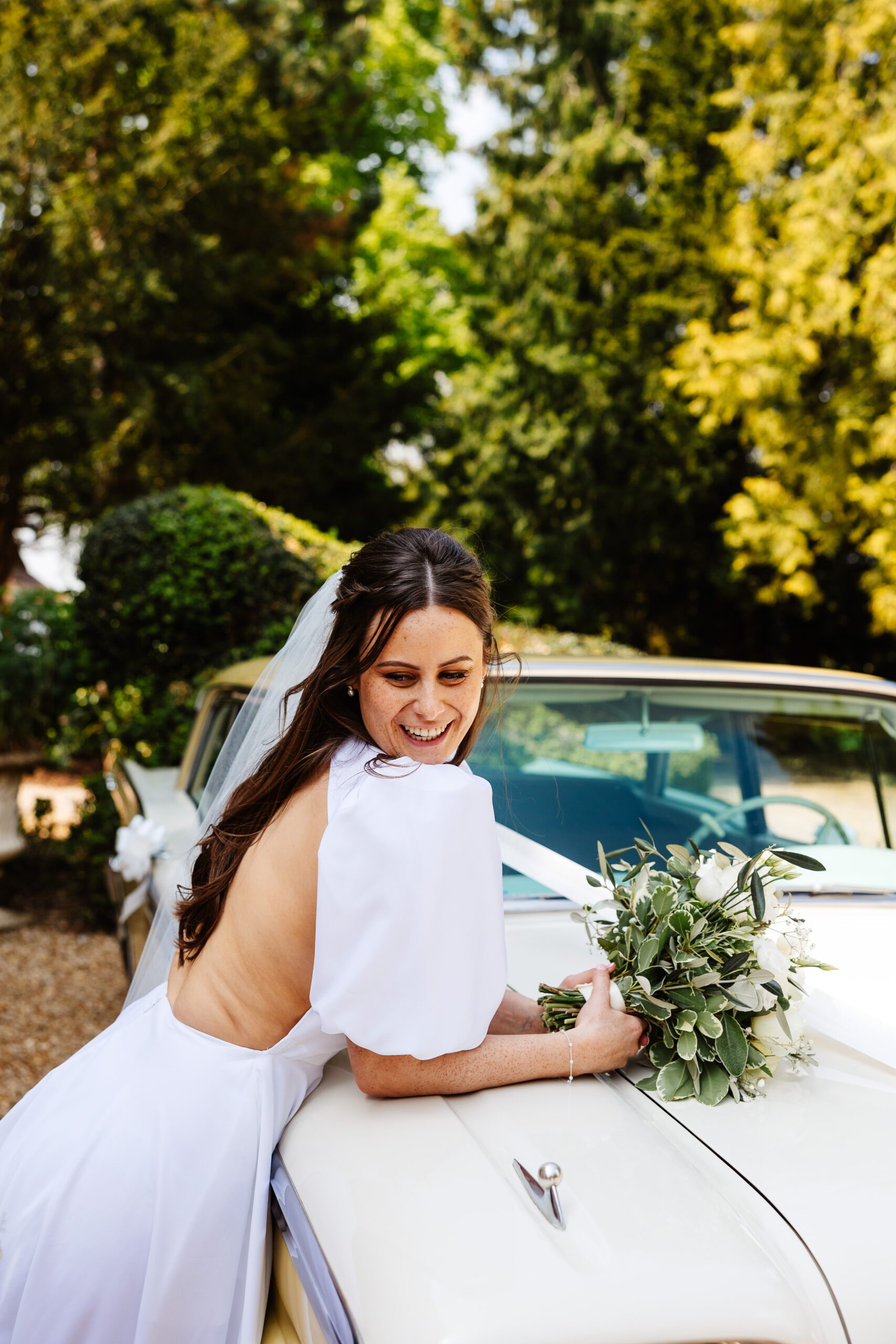 A bride in her wedding dress and white veil leaning over a car holding her flowers and smiling.