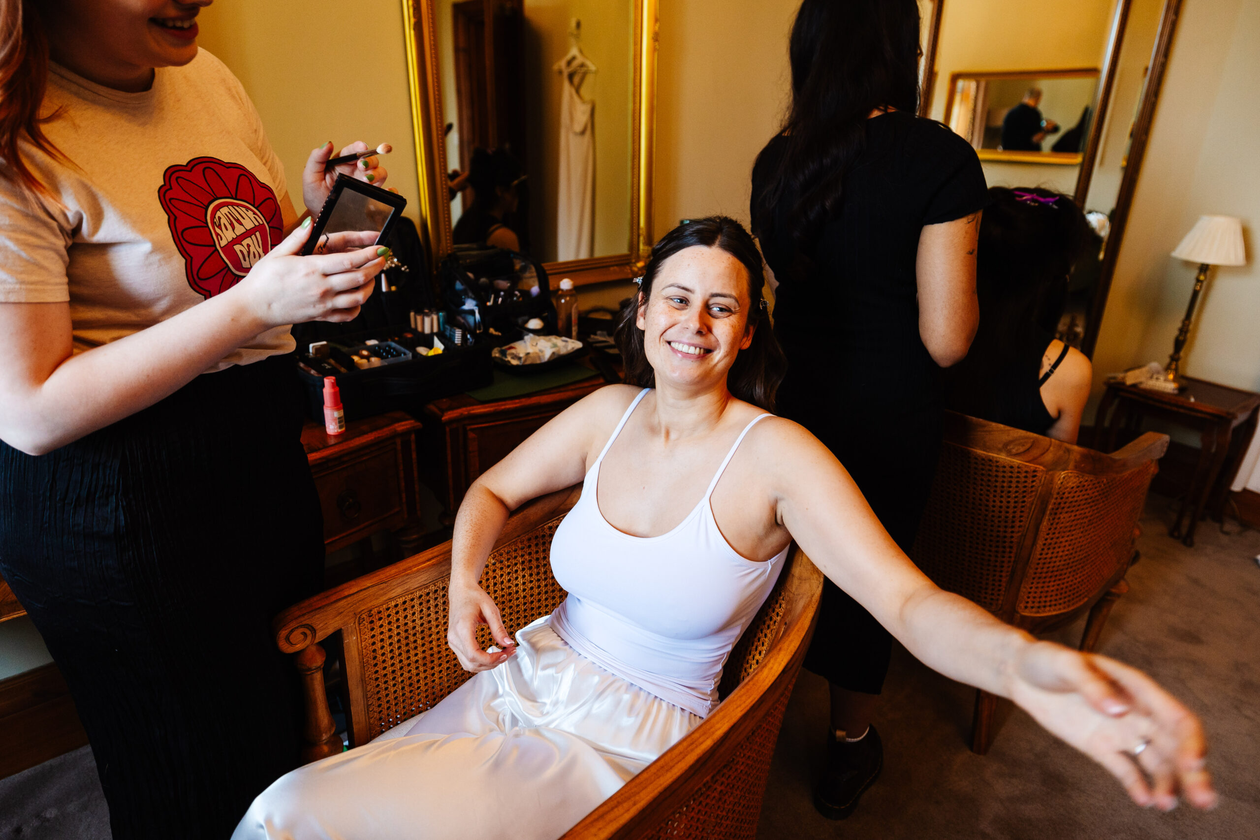 The bride sat in a chair in a white vest top and white silk bottoms having her make up done professionally. The bride has a big smile on her face.