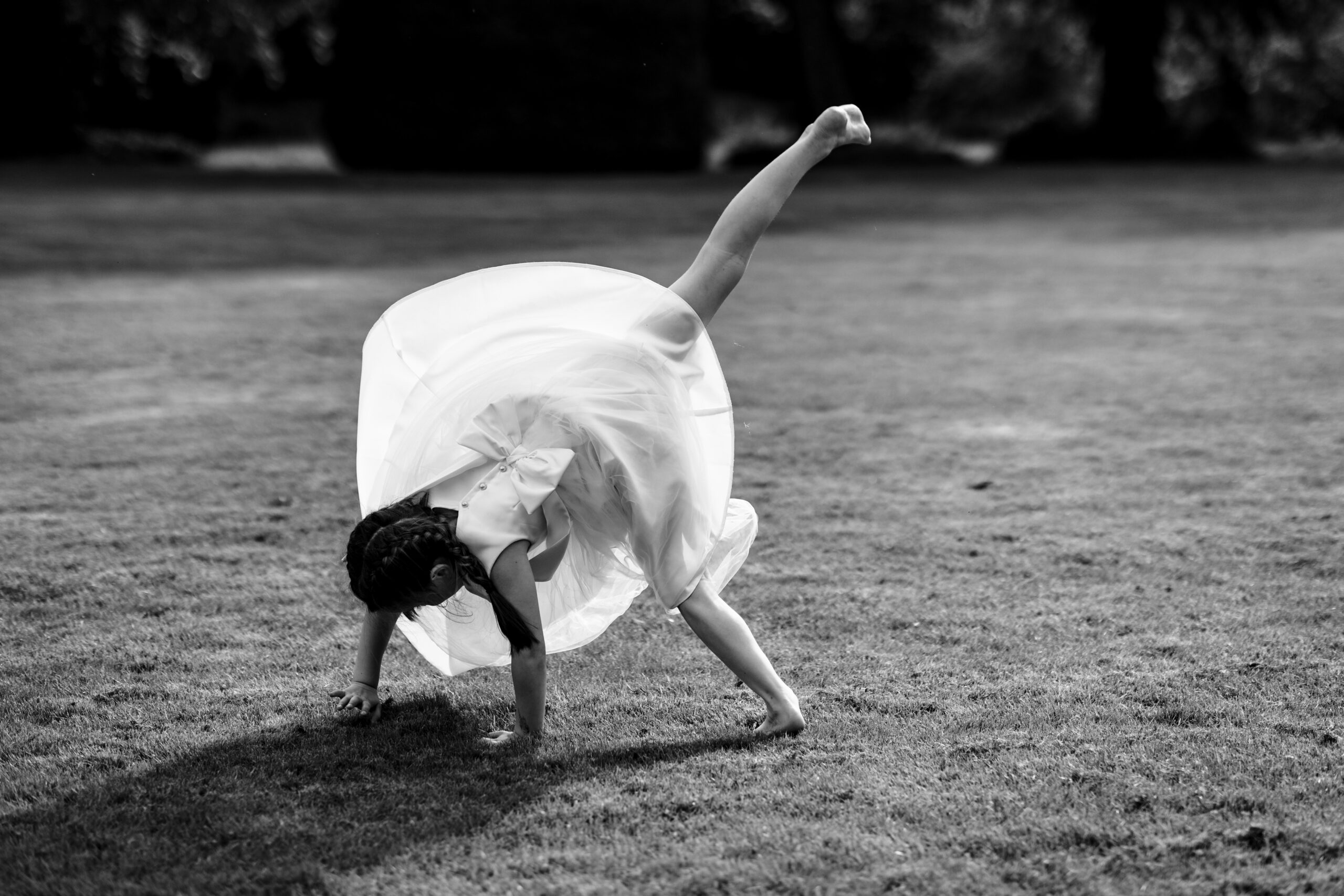 A little girl in a white dress doing a cartwheel on the grass. This image is black and white.