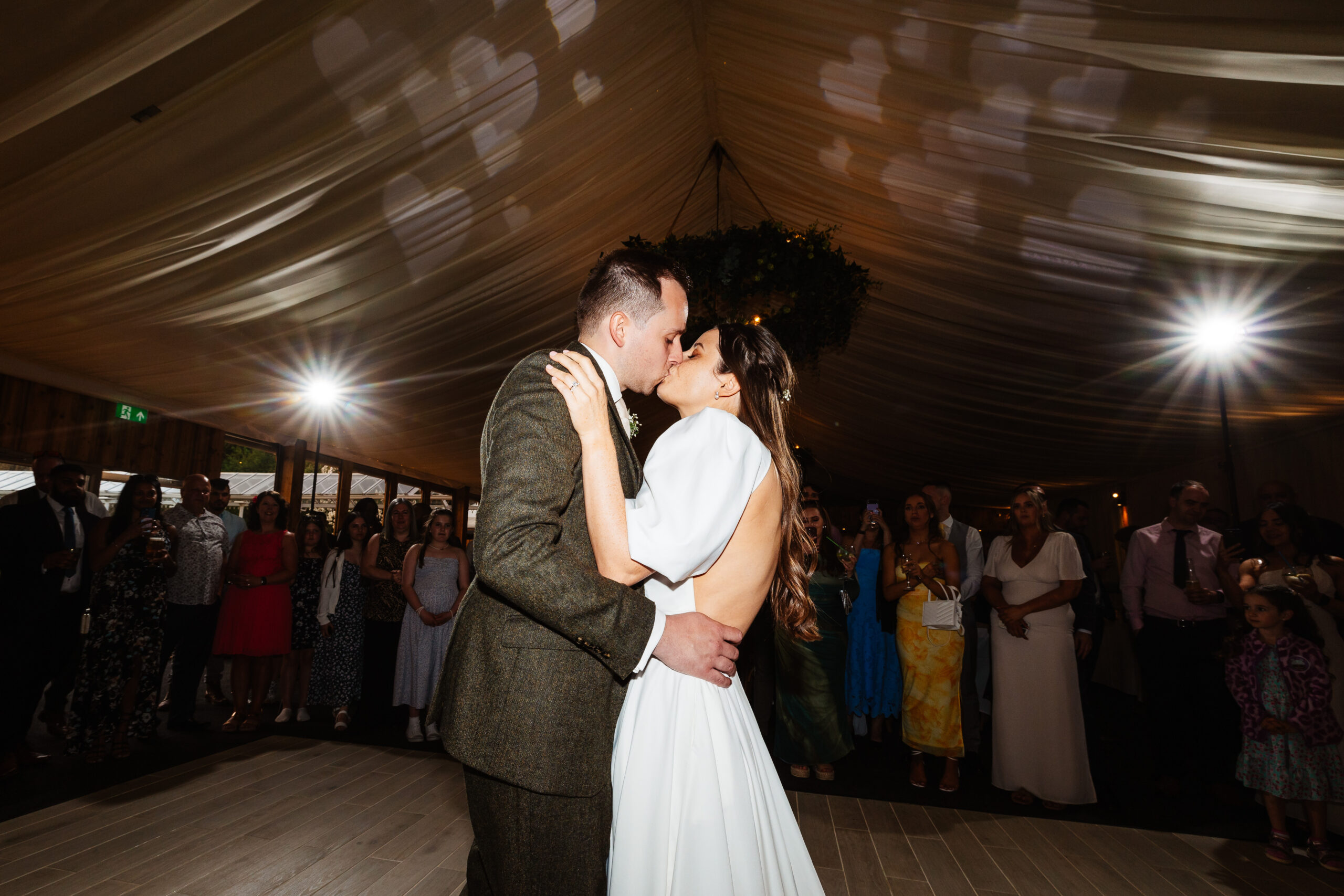 A bride and groom sharing a kiss during their first dance. Guests are surrounding them and the lights are shining in the background.