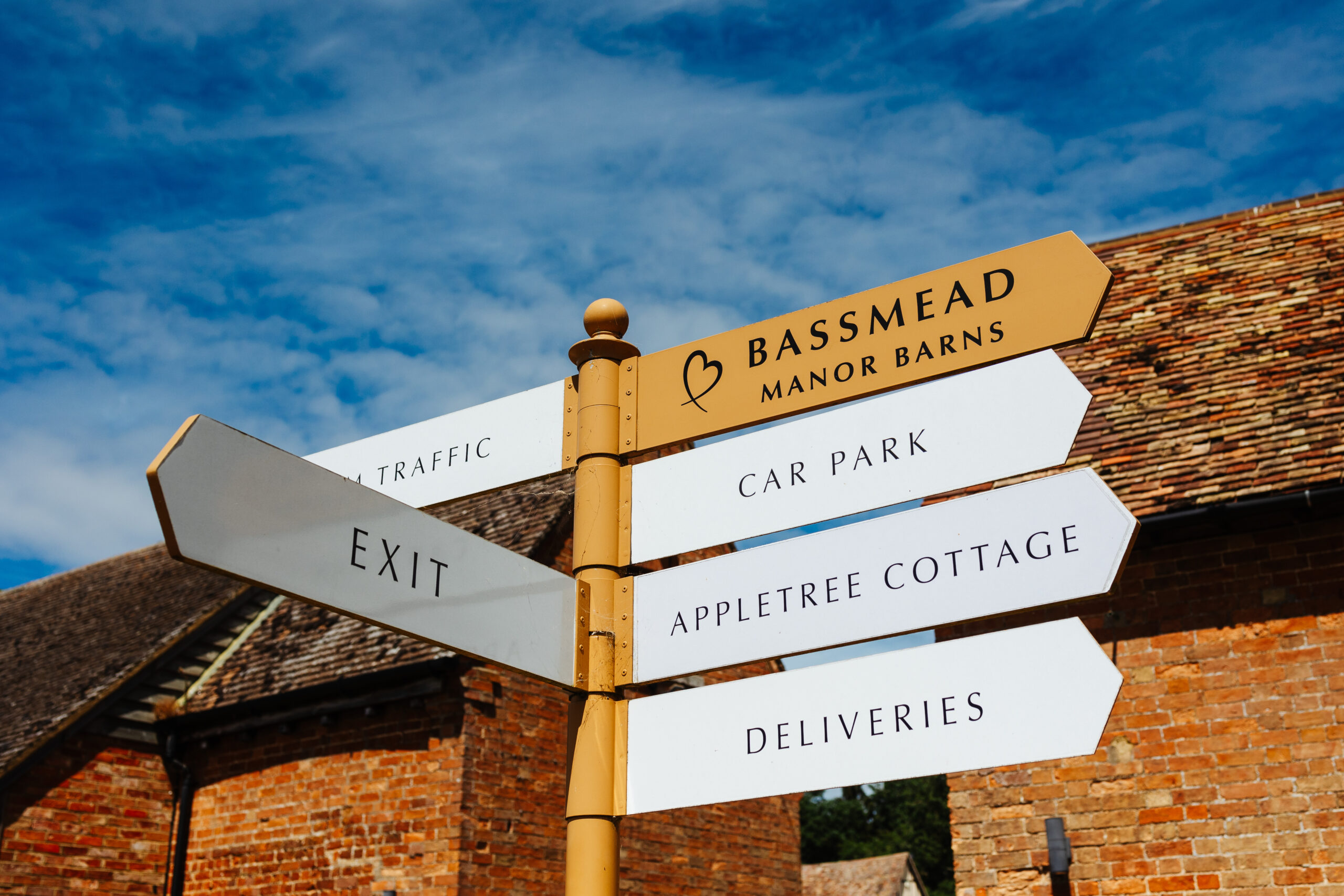 Signage for the venue of Bassmead Manor Barns. It is outside and the sky is bright blue with wispy white clouds.