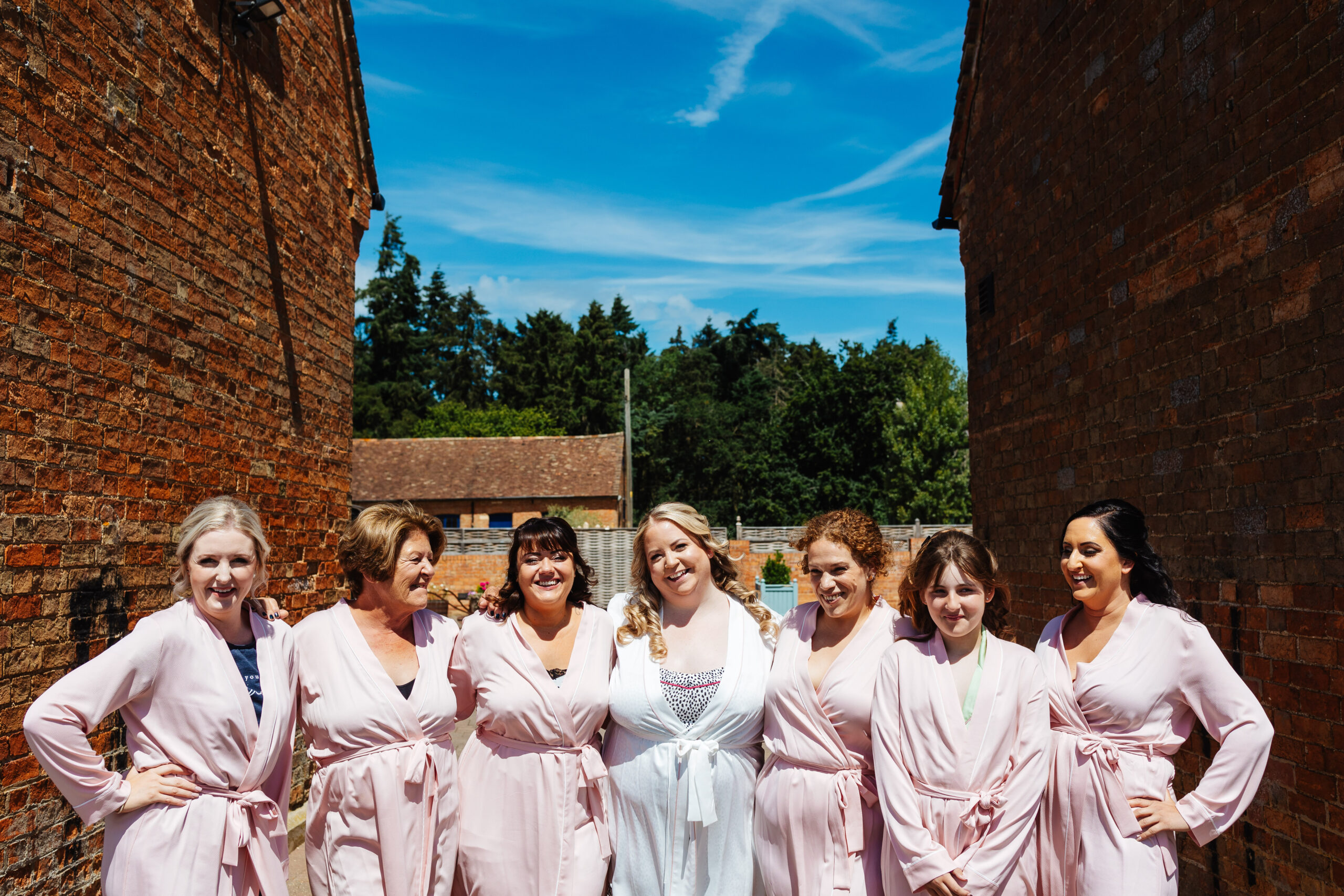 The bridal party stood outside. It is a sunny day and the sky is blue. They are smiling at the camera and are dressed in pink dressing gowns, except the bride who is in a white one.