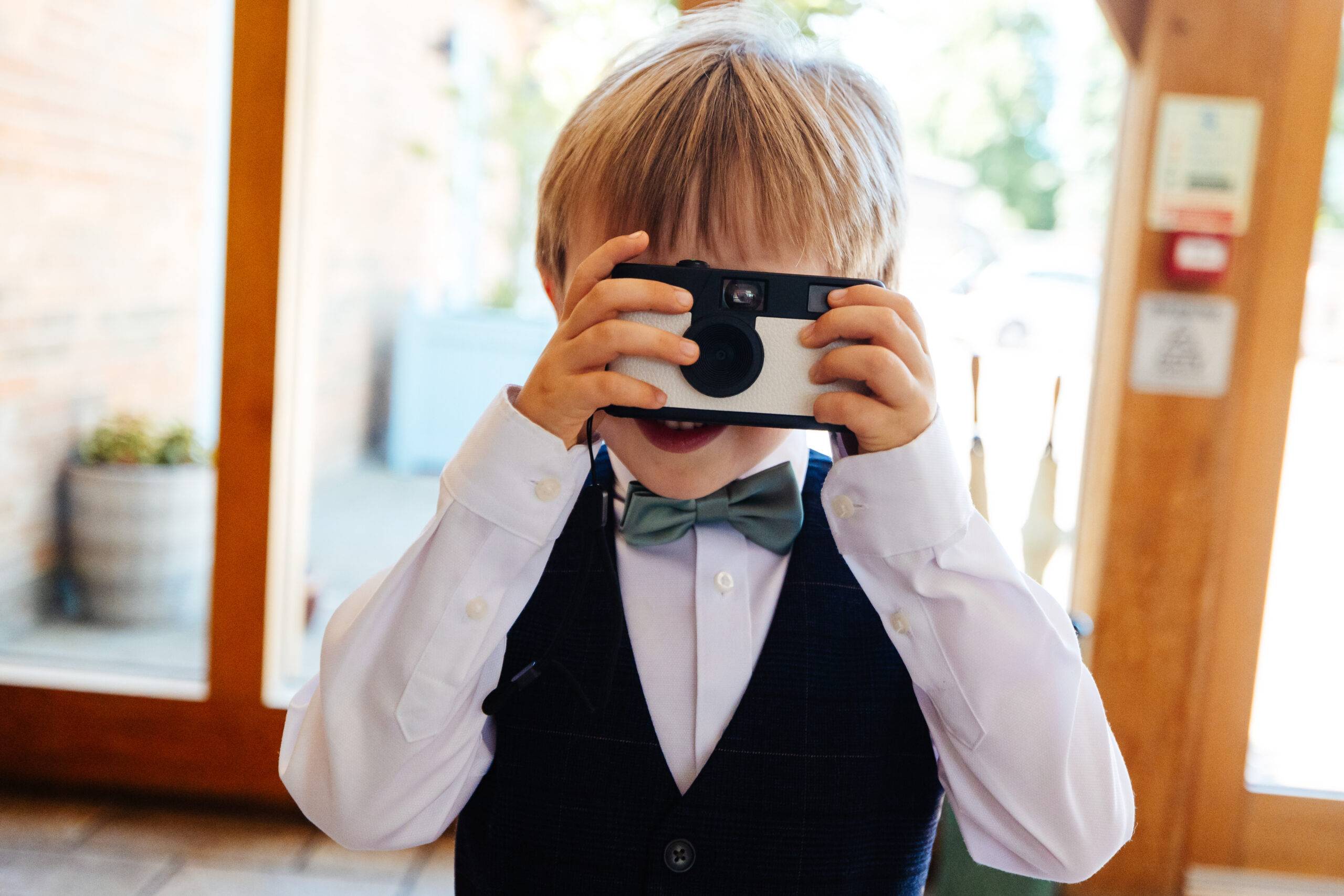 A little boy wearing a white shirt with a little turquoise bow tie. He is holding a disposable camera up to his face and has his finger on the button ready!