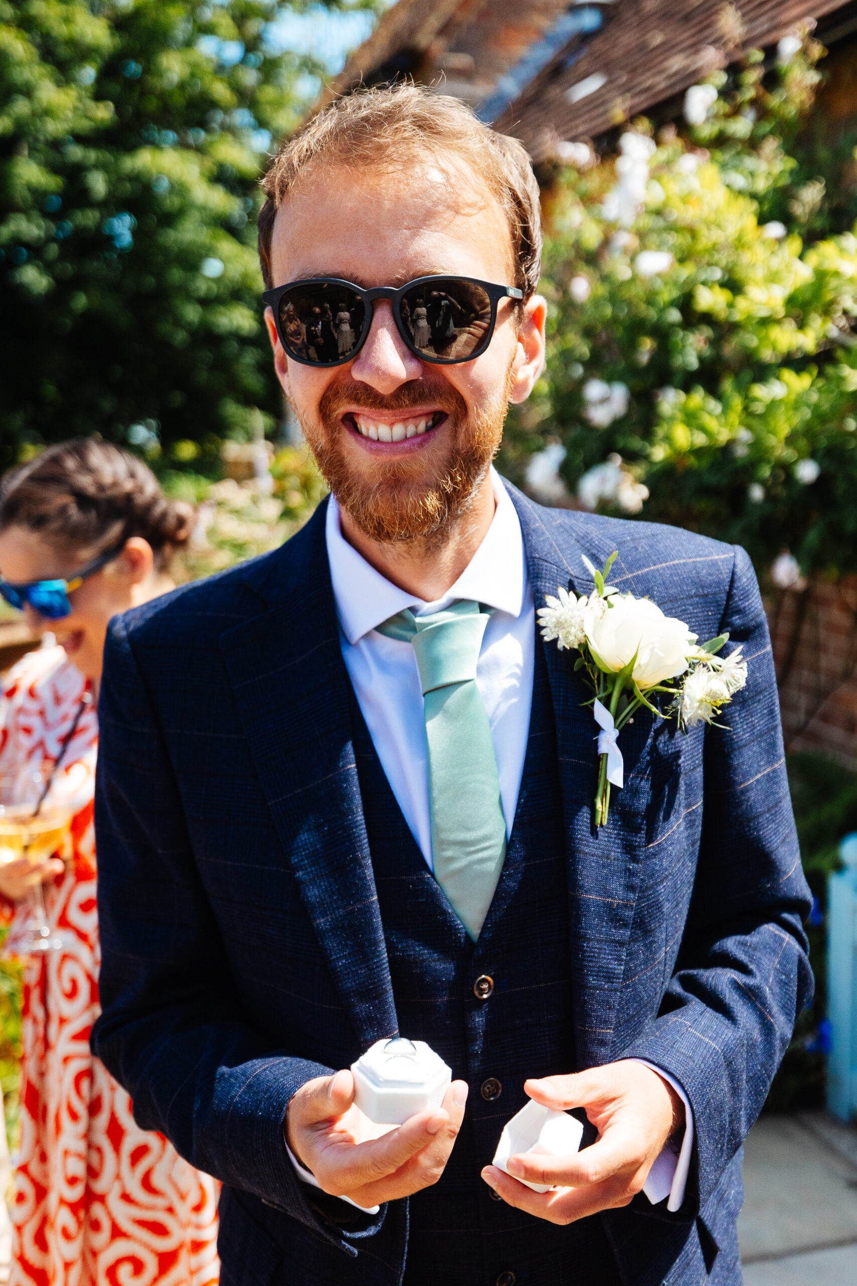 A photo of a man in a navy striped suit with a white shirt and pale green tie. He is wearing sunglasses and is smiling. He is holding the ring box.