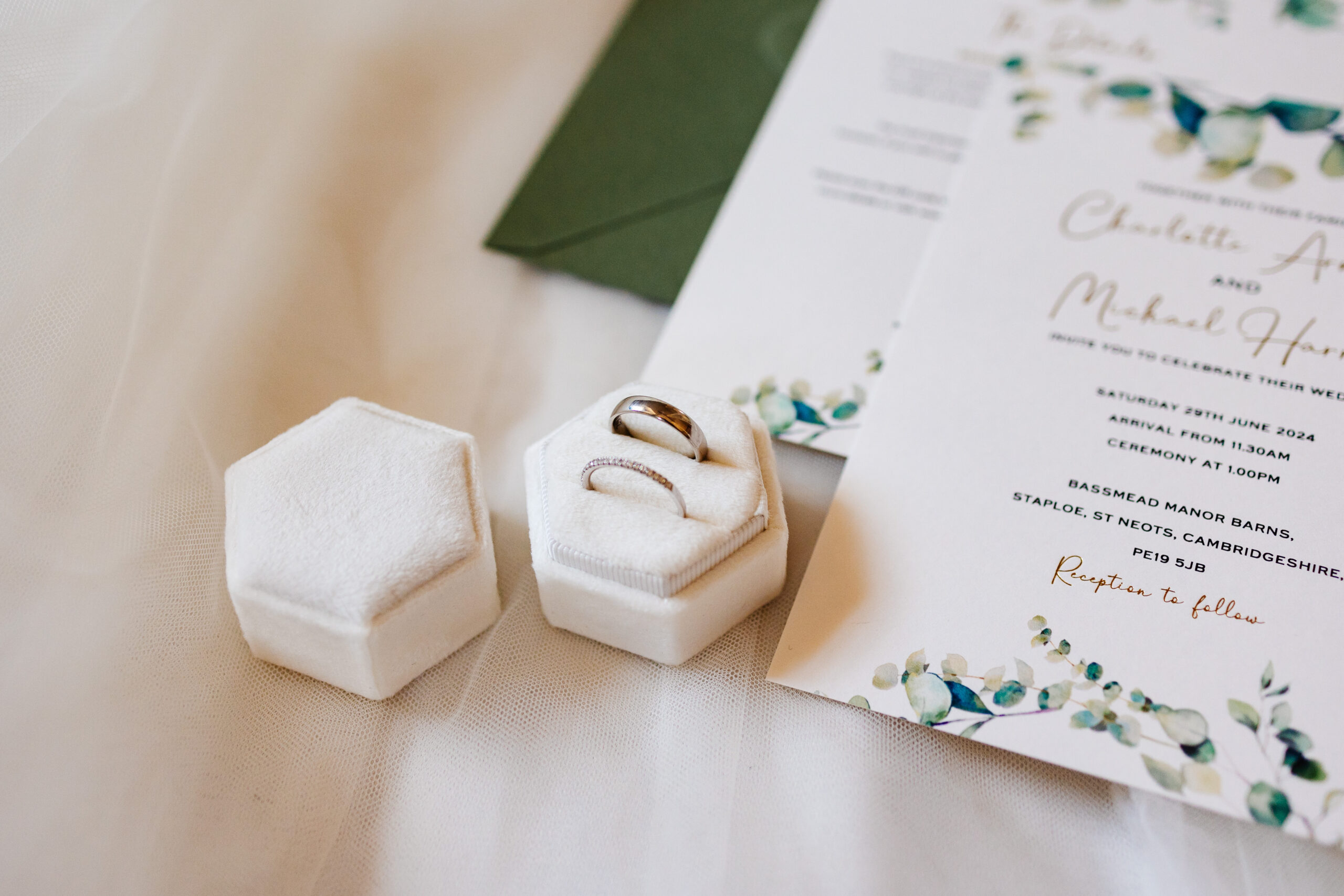 Charlotte & Michael's wedding invite which has curved writing on a white background and eucalyptus hand drawn on the bottom of it.
There is also a white hexagonal ring box containing two rings in it.