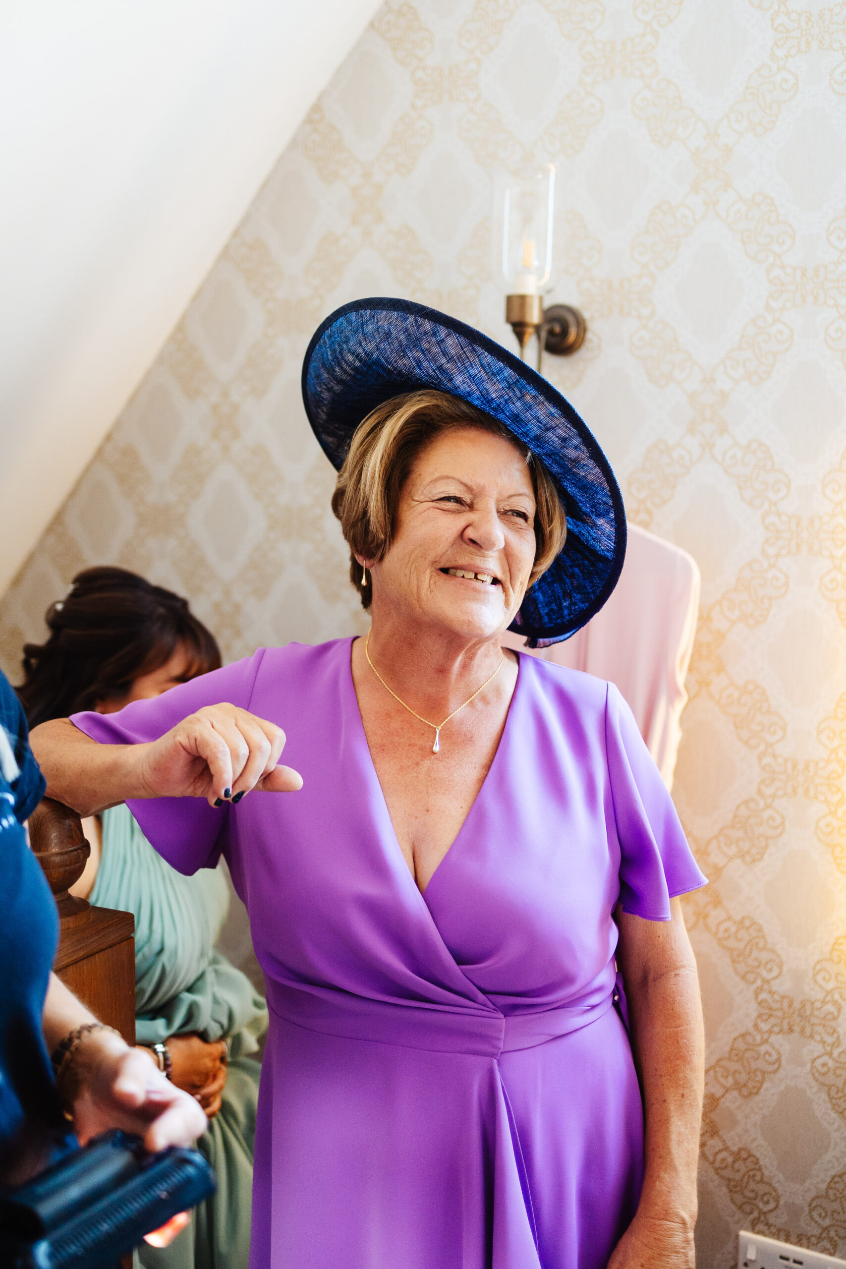 A photo of a lady in a bright purple dress with a blue hat on. She has a delicate necklace with a pearl drop in the middle. She is smiling at the bride.