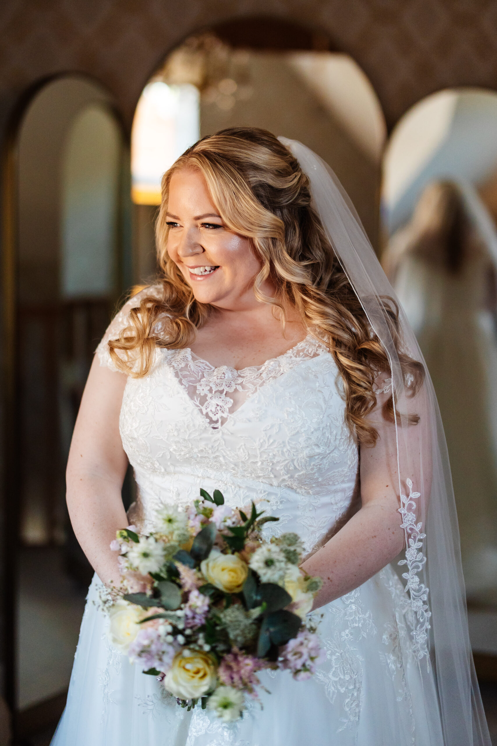 The bride in her lacey white dress and long flowing veil. She is holding her bouquet of flowers and is smiling at her family who you can't see (off screen).