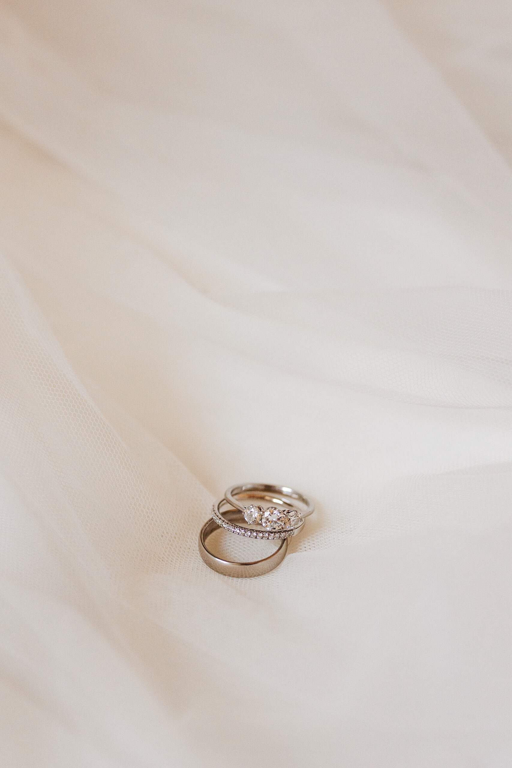 Three silver rings; one is a plain wedding band, one is a band with diamantes on and one has three diamonds on. They are resting on the bride's veil