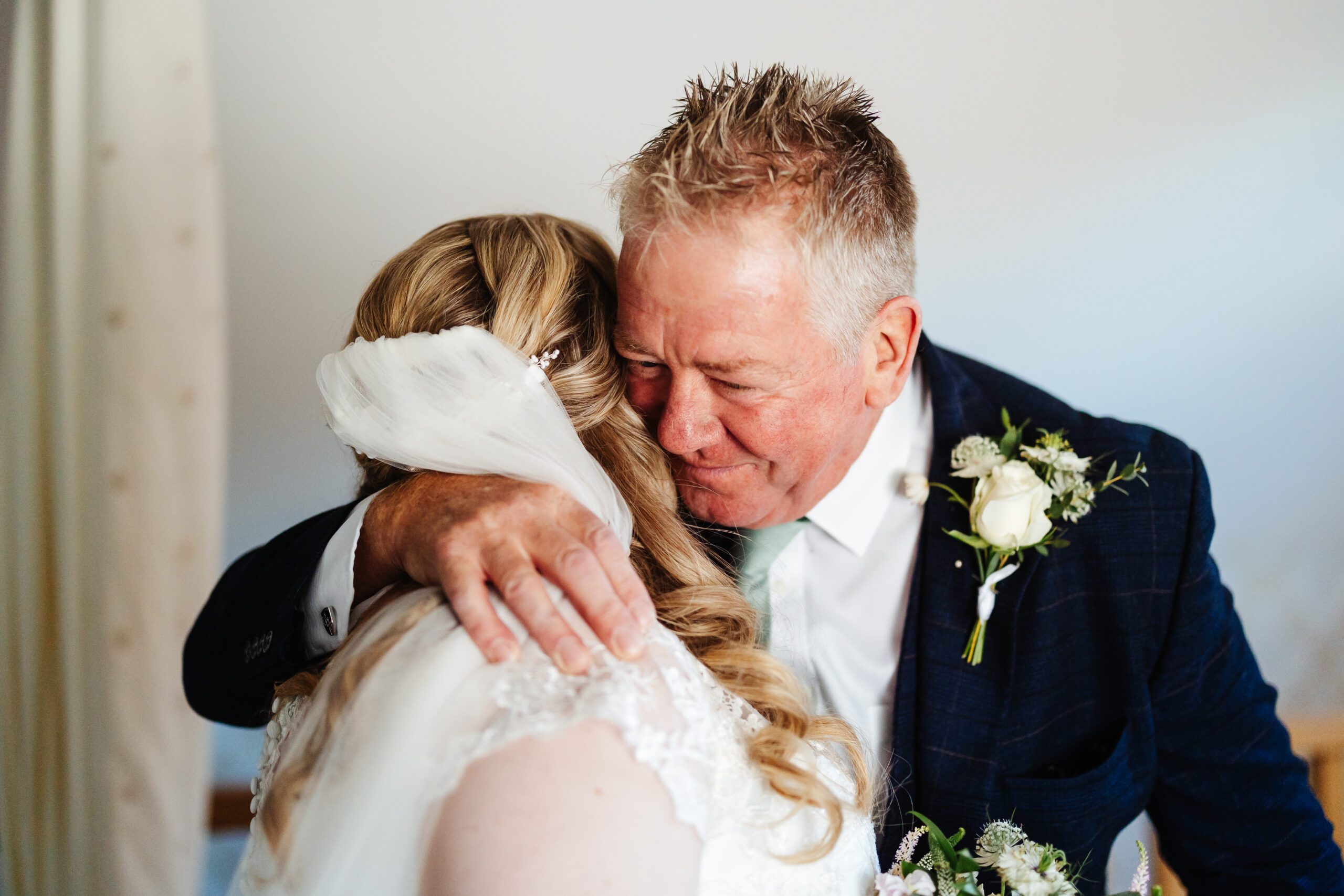 A bride hugging her father. He is wearing a navy suit and has one arm around her. He looks very happy.