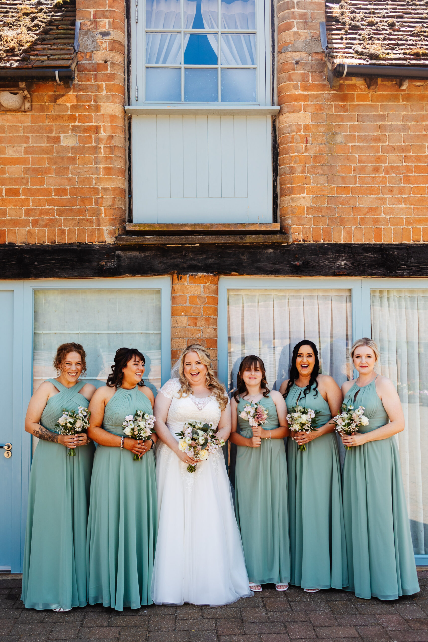 A portait image of the bridal party in their dresses and all glammed up. They are wearing a variety of styles of dresses in turquoise and have little bouquets of flowers to match the bride. They are all smiling and looking at the camera.