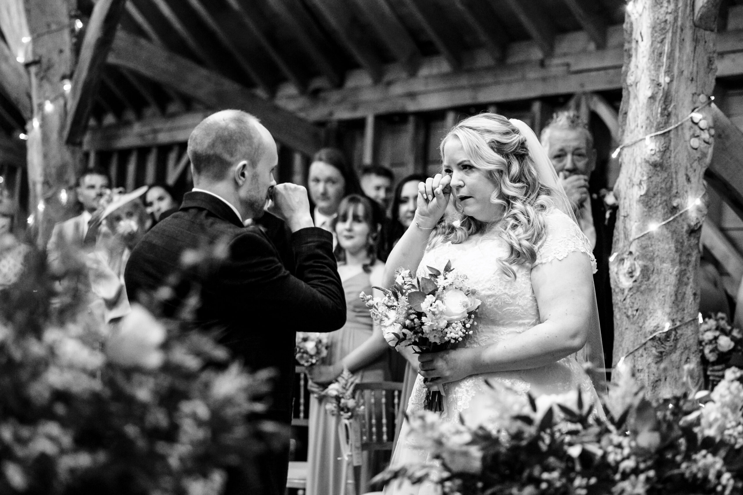 A black and white image of the bride and groom. They are both wiping away tears. The father in the background has his hand to his mouth in a very emotional way too. 